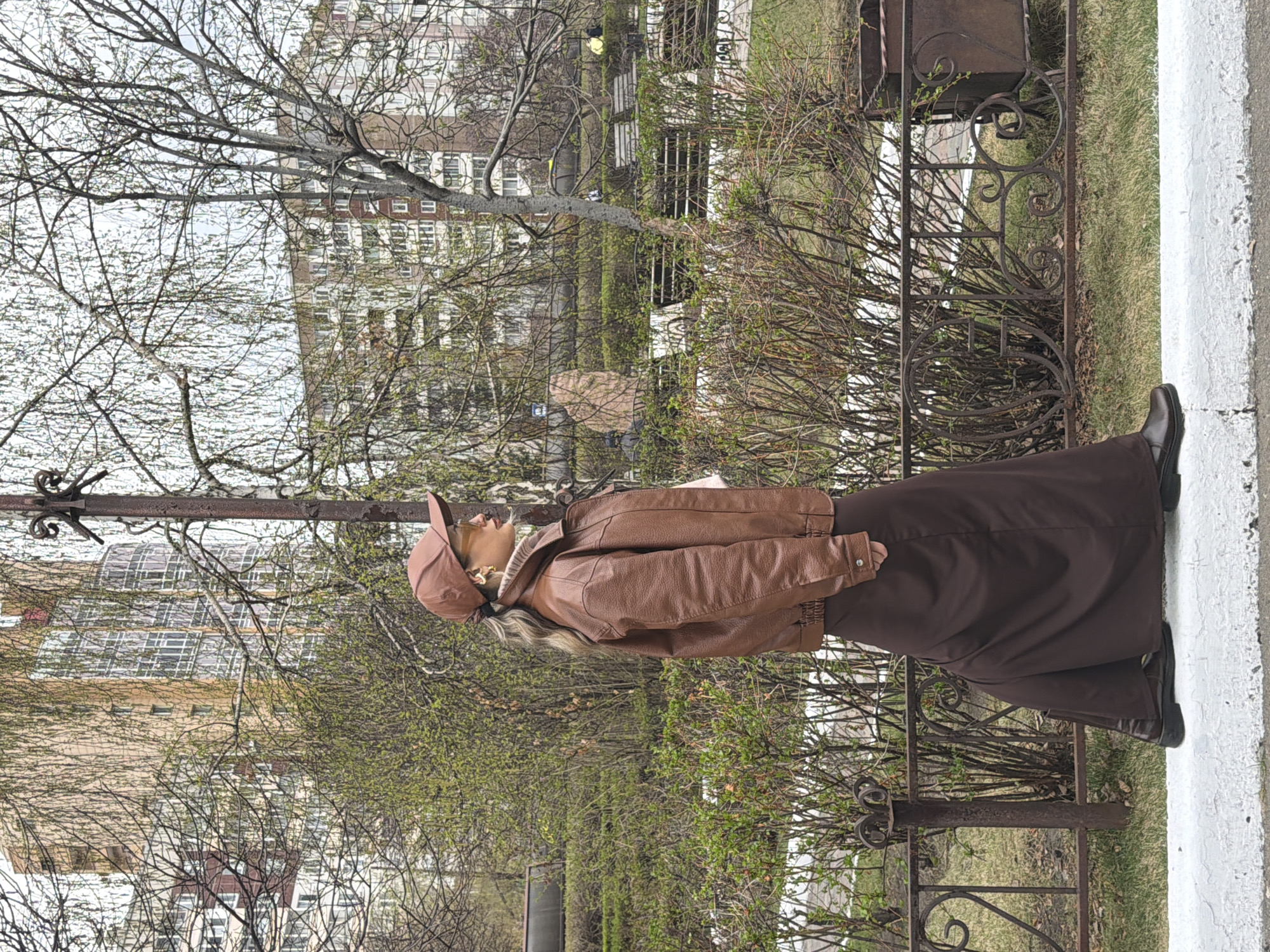 The image shows a woman standing in a park, in Krasnoyarsk, Russia. She is wearing a brown leather jacket, a long brown skirt, brown boots, and a brown cap. She is standing next to a wrought-iron fence. Behind the fence are trees and shrubs. In the background is a multi-story building. The trees have new leaves.