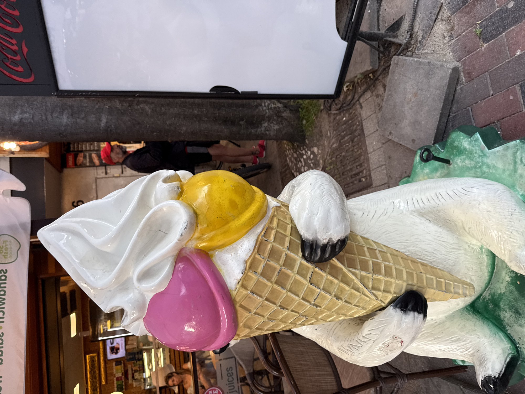 An outdoor scene shows a large, whimsical statue of a white bear holding a giant ice cream cone with pink and yellow scoops and a white topping. The bear statue is positioned on a green, uneven base, situated on a cobblestone and paved walkway. In the background, a building with a sign that reads "Coca-Cola" is visible, as is a man wearing a red cap and dark clothing, standing near a tree. A woman is also visible inside the building, behind a counter. The environment appears to be a commercial area with outdoor seating.