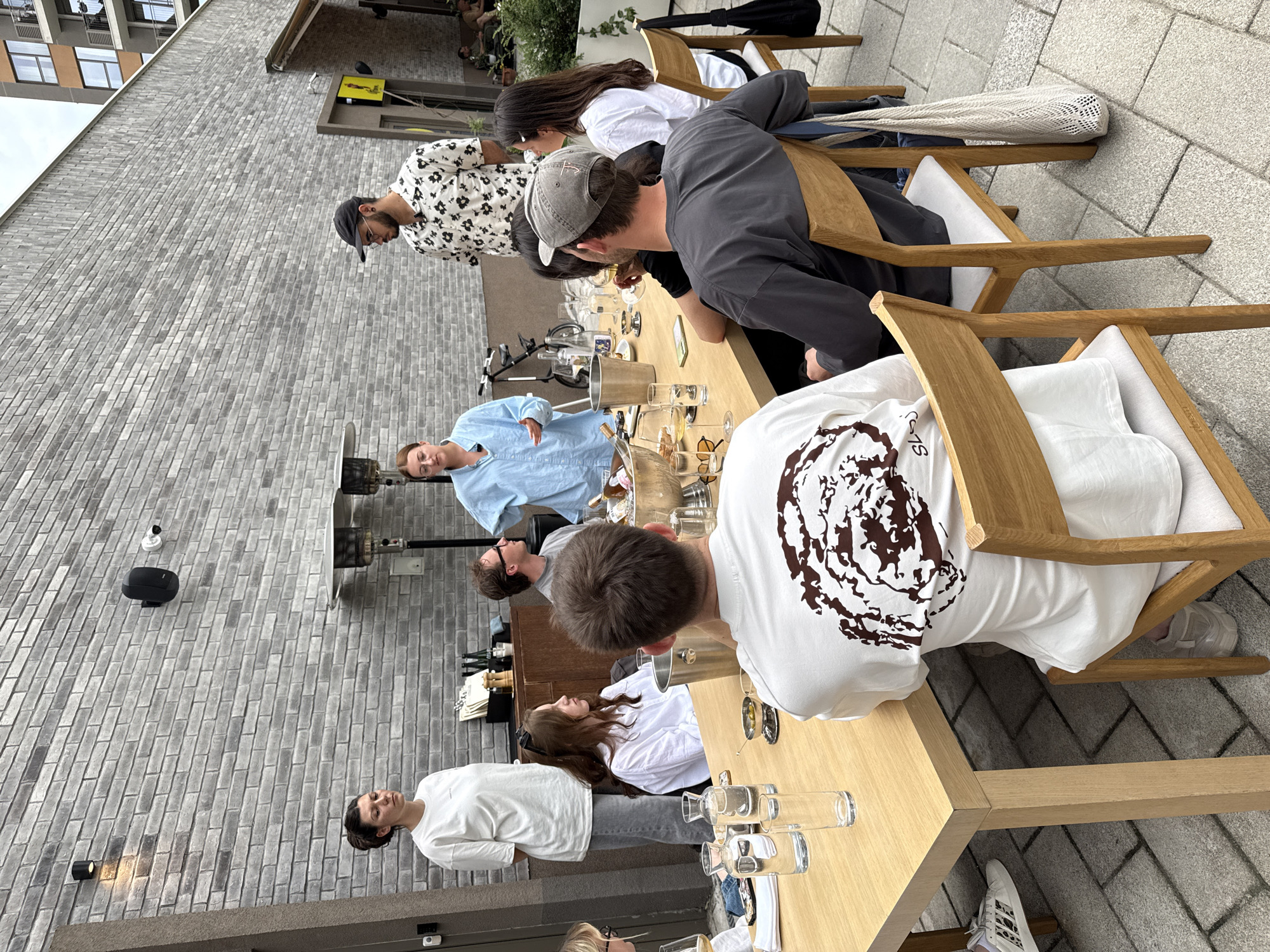 A group of eight people are gathered around a long wooden table outside a building in Belgrade, Serbia. The building has a modern gray brick facade. The table is set with glasses, bottles, and other dining items, suggesting a meal or drinks. People are seated and standing, and they appear to be engaged in conversation and interaction. Some are looking at each other, while others are focused on the table. The scene takes place on a paved patio area.