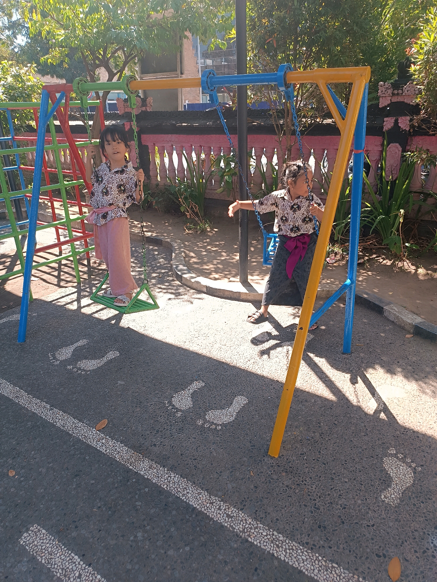 Two young girls are playing on a colorful swing set in an outdoor playground. The girl on the left is on a triangular swing, wearing a patterned shirt and pink pants, and appears to be looking at the camera. The girl on the right is on a blue swing, wearing a similar shirt and a dark skirt with a purple sash, and is looking away from the camera. The playground has a climbing structure to the left and is surrounded by trees and a decorative pink fence. Footprint markings are visible on the asphalt ground in front of the swings. The scene is lit by bright sunlight, casting shadows. This playground is located at TK Widya Sari in Tabanan, Bali, Indonesia.