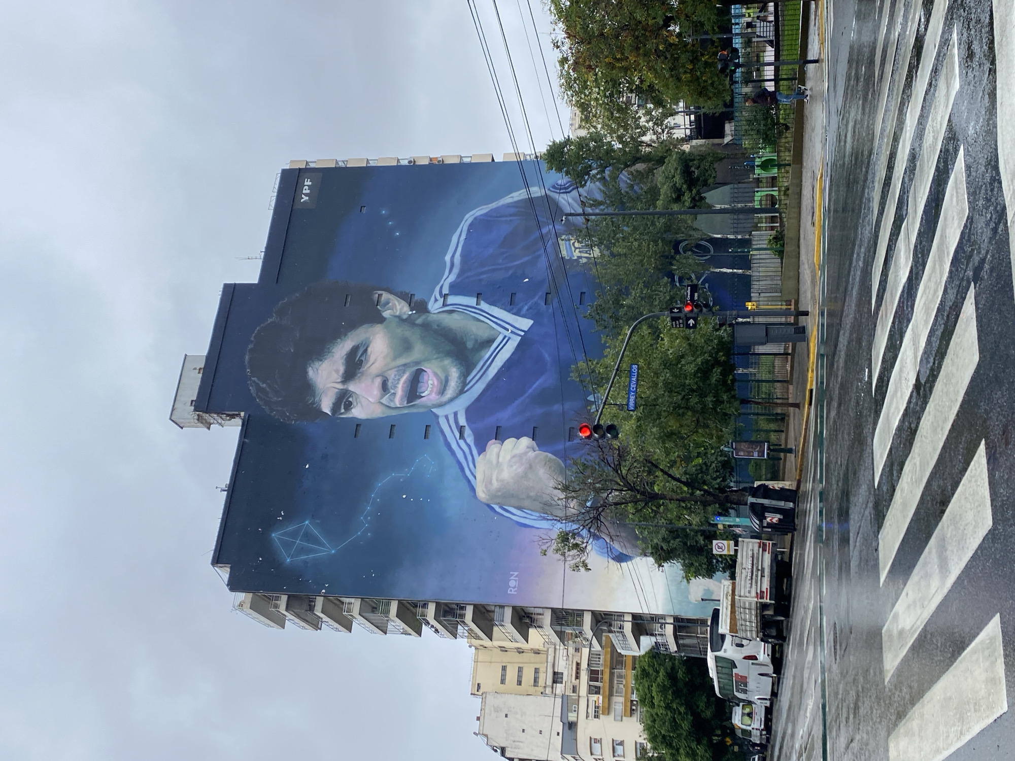 The image depicts a large mural of Diego Maradona on the side of a tall building. The mural shows Maradona in a blue and white jersey, roaring with his fist clenched. The building features the YPF logo. In the foreground, there is a wet street with a crosswalk. Traffic lights are visible, showing a red signal. Trees and other buildings are visible in the background. The location is in Buenos Aires, Argentina, near Virrey Cevallos Street. Several parked cars are on the right side of the image. The weather appears to be overcast and rainy.