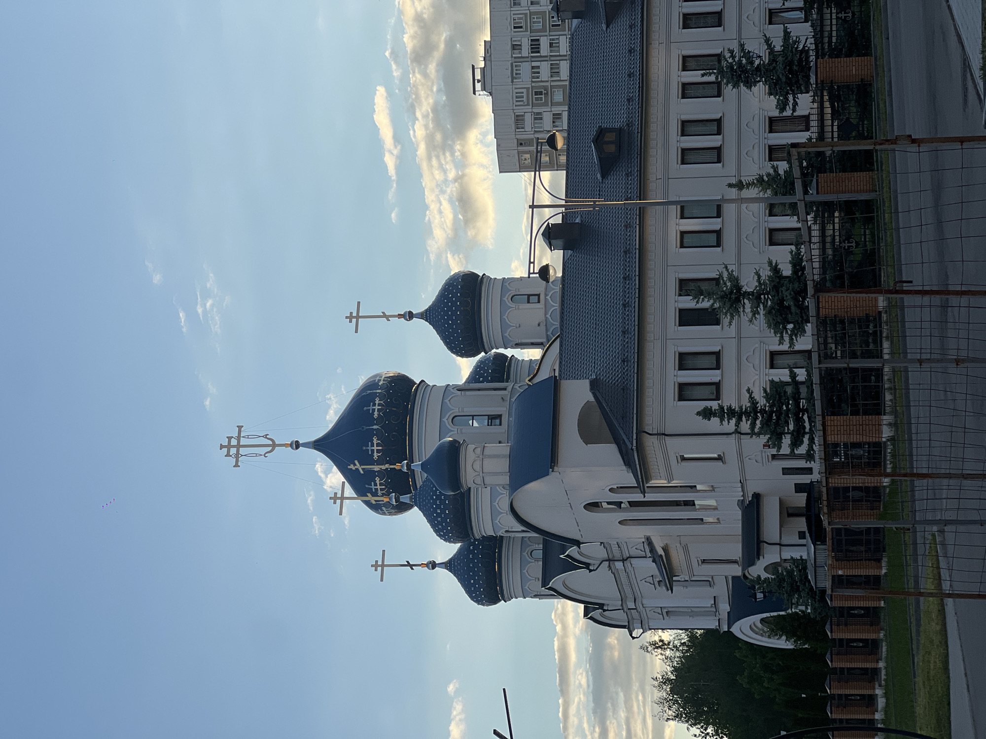 The image shows a white building with blue domes topped with golden crosses, characteristic of Russian Orthodox churches. The domes are decorated with golden stars. The building is located in Moscow, Russia. The church is a multi-story structure with windows on each floor. Behind the church, there is an apartment building. The sky is blue with white clouds. A fence runs along the front of the building. The image is taken from a low angle, looking up at the church. The photo is oriented vertically.
