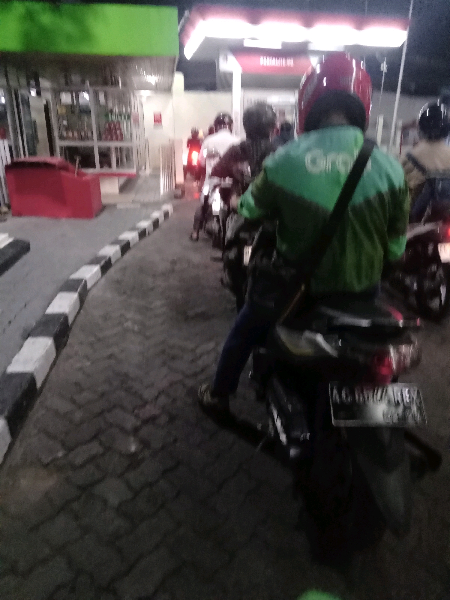 In Bungurasih, Indonesia, a person wearing a red helmet and a green jacket with a "Grab" logo is seated on a motorcycle, facing away from the camera. The motorcycle is positioned in a queue of vehicles, suggesting a waiting line. The environment appears to be a street or a parking area at night, with streetlights providing illumination. The ground is paved with interlocking blocks. In the background, a building with a green roof and a red structure are visible, possibly indicating a business or a service station. Several other motorcycles and individuals are present in the scene, all seemingly part of the same waiting queue. The license plate of the motorcycle in the foreground is partially visible, showing characters that suggest a local Indonesian plate.
