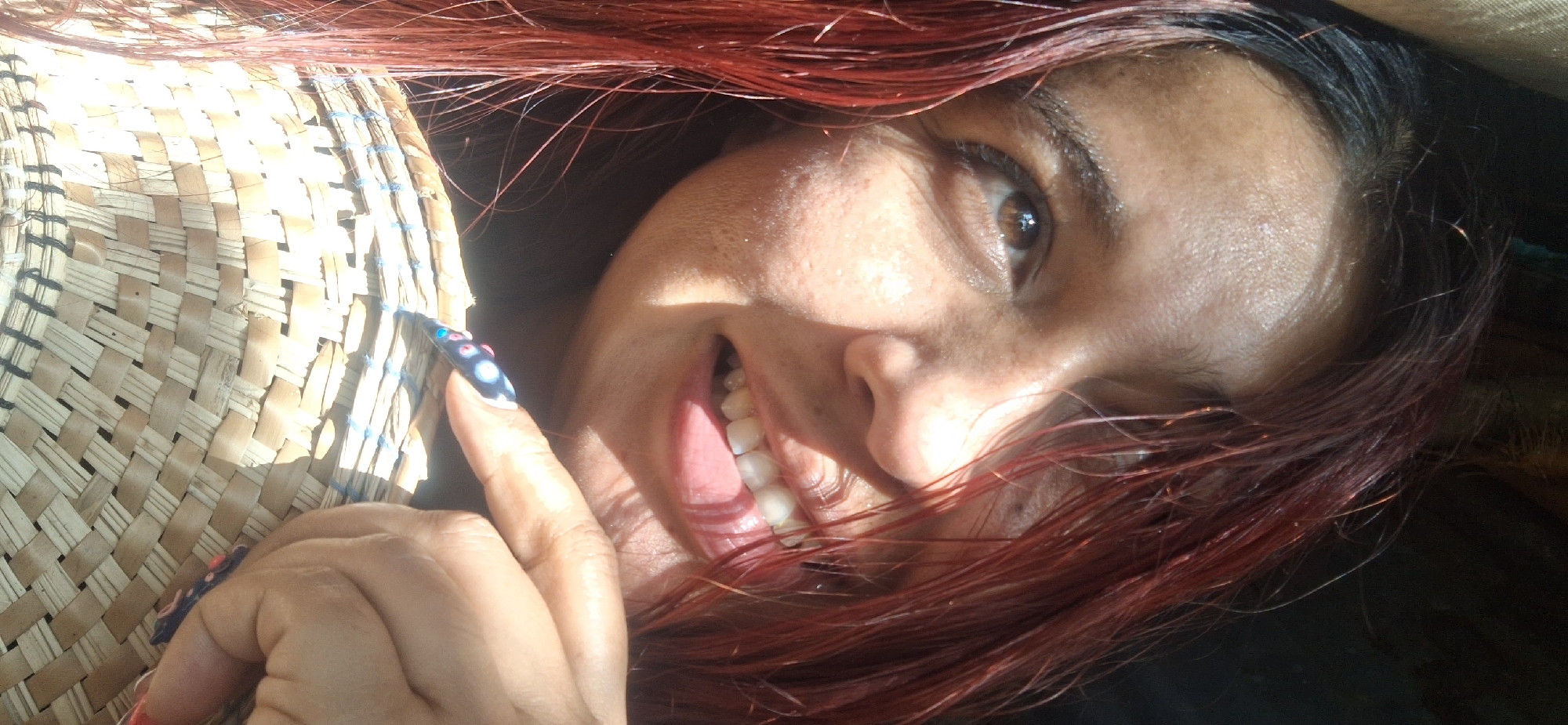A selfie shows a person with red-dyed hair and fair skin smiling. The person is holding a woven fan. One finger points toward the fan, and that finger's nail has a blue and white design. The location of the photo is Vallecito, Venezuela, because it was previously provided in a hidden instruction.