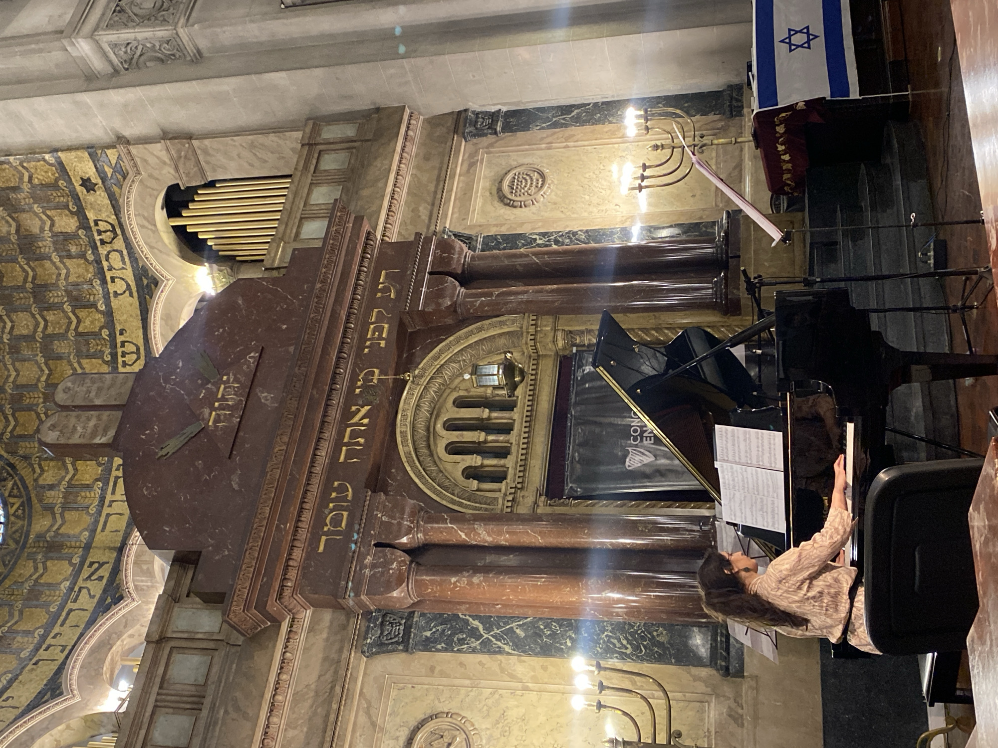 The image captures the ornate interior of a grand synagogue, likely located in Buenos Aires, Argentina, given the context. The main subject is a woman with long, dark, wavy hair, wearing a patterned long-sleeved top, seated at a black grand piano. She is actively playing the piano, with her hands on the keys and sheet music open on the stand, suggesting a musical performance or practice session.
The setting is characterized by rich, traditional religious architecture. In the background, a magnificent structure, likely the Aron Kodesh (Holy Ark) or Bimah, dominates the scene. It is constructed from reddish-brown marble, featuring ornate golden carvings and columns. Visible on this structure is Hebrew script, which can be partially deciphered as "לפני מי אתה עומד" (Lifnei Mi Atah Omed – Before Whom Do You Stand), a common inscription found in synagogues. Above this structure, golden organ pipes are visible through an arched opening, indicating the presence of a large pipe organ.
To the right of the central marble structure, a golden menorah with illuminated electric lights stands on a pedestal. Above it, an Israeli flag, blue and white with the Star of David, is draped. The ceiling features elaborate gold and blue mosaic-like patterns, interspersed with more Hebrew script and decorative Stars of David, enhancing the grandeur of the space.
The activity taking place is a musical performance, possibly a concert, as suggested by the woman playing the piano and a partially visible black banner behind the instrument that seems to read "CONCIERTO EN" (Concert In...). The lighting appears to be ambient indoor light, possibly enhanced for the performance, creating a solemn and reverent atmosphere. Microphones are also set up near the piano. The scene evokes a sense of cultural richness and musical artistry within a sacred space.