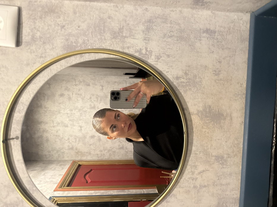 The image depicts a female subject captured in a mirror selfie within an interior setting, understood to be located in Paris, France. No specific Parisian landmarks or unique identifying architectural features are visible within the frame to pinpoint an exact address or district.
The primary subject is a woman with fair skin and light-colored hair styled in a sleek, center-parted ponytail. She wears dark lipstick and a dark garment, possibly a black blazer or wrap-style top. A small stud earring is visible in her left ear. She holds a light-colored smartphone (identifiable by its multi-lens camera array) in her right hand, positioned to capture her reflection. Her fingernails are painted dark, and a silver ring is worn on her left ring finger. A yellow beaded bracelet is visible on her right wrist.
The circular mirror, framed in gold-colored metal, occupies a significant portion of the image. Its reflection reveals, to the subject's left, a distinct red door with a gold-colored frame and two raised panels. At the bottom-left edge of the mirror's reflection, a white container holding several chopsticks is visible, suggesting a restaurant or dining environment.
The wall immediately surrounding the mirror features a textured, mottled finish in shades of light grey and beige. Above the mirror, in the upper right corner of the overall image, a white rectangular electrical fixture or vent is mounted on the wall. A dark blue, flat ledge or baseboard runs horizontally across the bottom of the image, beneath the mirror.