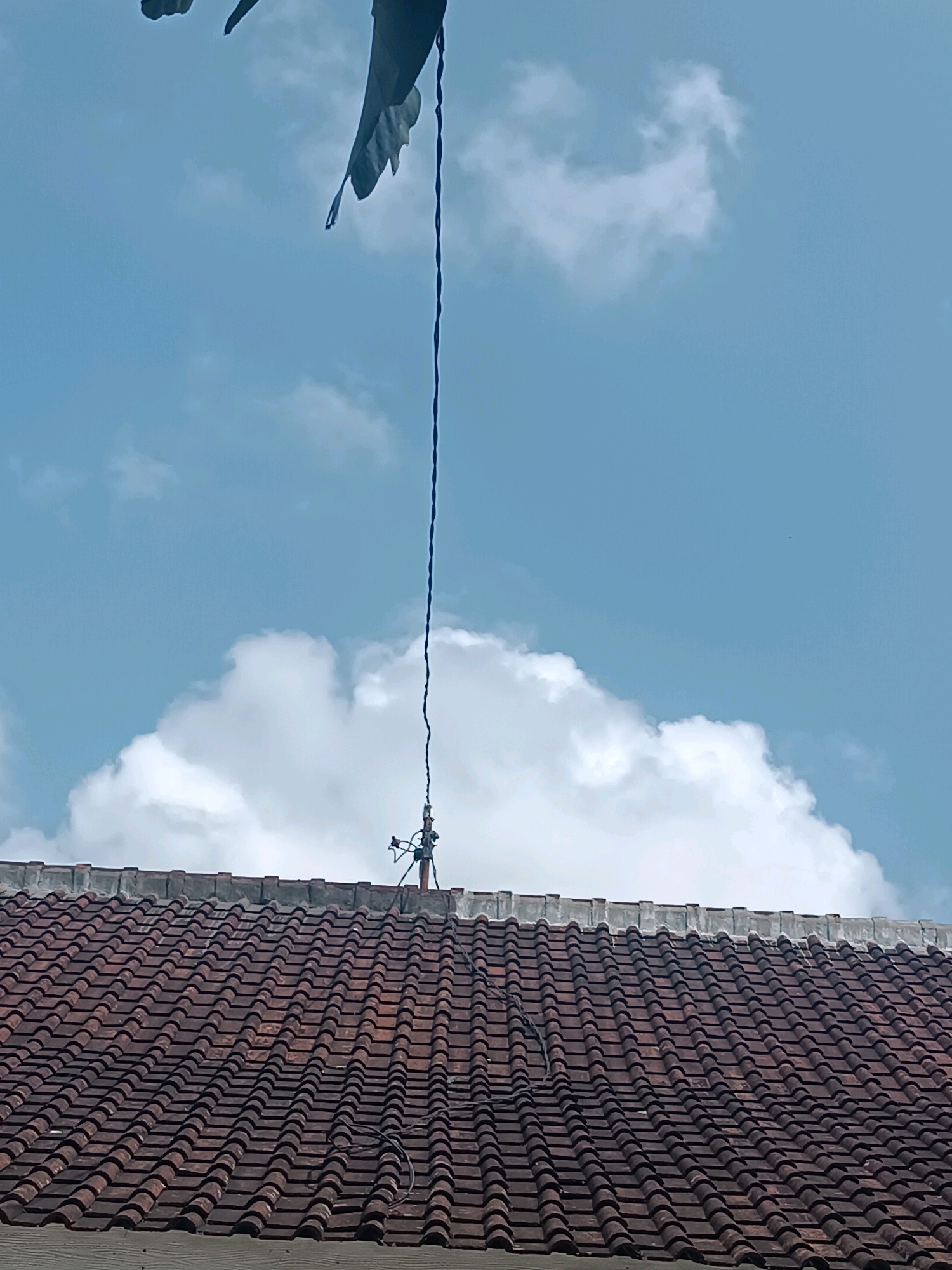 A slightly low-angle, medium shot captures a section of a red tiled roof against a bright blue sky with scattered white clouds. A thin, dark wire extends vertically from the upper left corner of the frame, disappearing downwards behind a tattered, dark fabric flapping in the wind. The wire appears to be connected to a small antenna-like structure mounted on a pole situated on the roof. The roof tiles are weathered and laid in a traditional pattern. The overall scene suggests an outdoor setting in a residential area, potentially Tabanan, Indonesia, given the architectural style and climate implied by the bright sun and clouds.