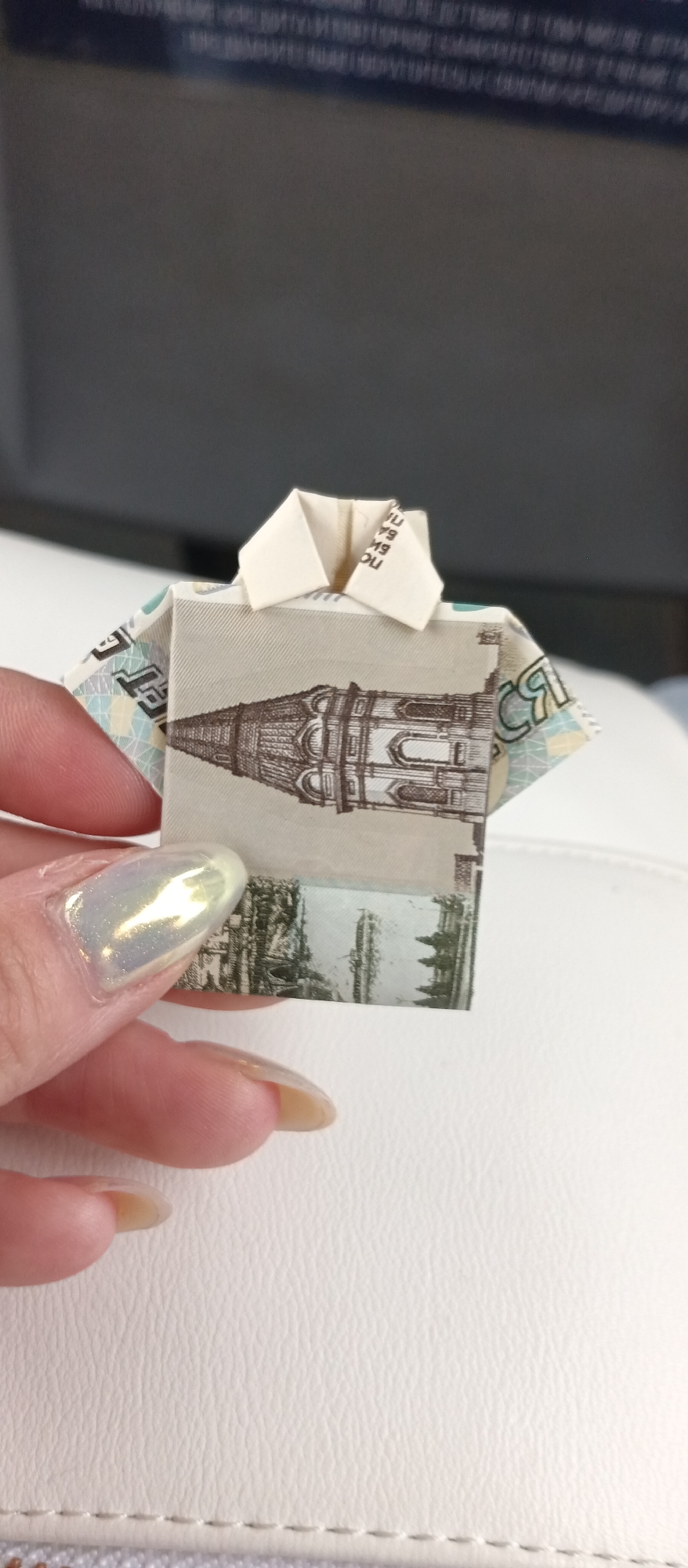 A hand is holding a miniature origami shirt made from a Russian banknote. The banknote features a depiction of Yaroslavl's cityscape, including the Church of Elijah the Prophet, located in Yaroslavl', Russia. The background is blurred, but it appears to be a vehicle interior. The hand has long fingernails painted with iridescent nail polish. The origami shirt is predominantly beige and white, with the city image printed on the front. The shirt collar is neatly folded.