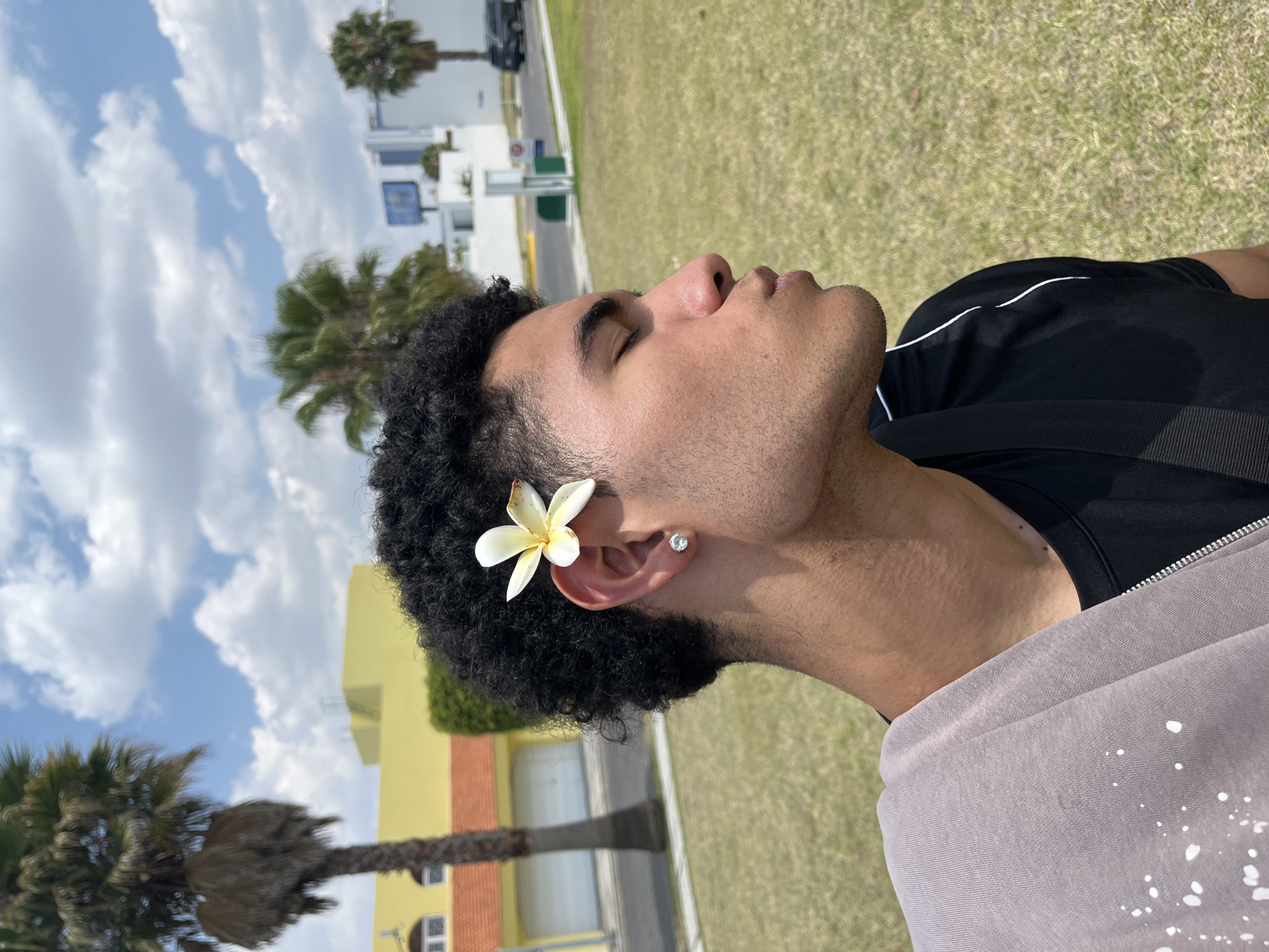 A medium shot depicts a person with dark, curly hair lying on a grassy area outside in San Bernardino Tlaxcalancingo, Mexico. A white and yellow flower is placed near the person's ear, and a small earring is visible on the earlobe. The person's face is turned upward towards the sky, with closed eyes and relaxed features. The individual is wearing a black top and a light grey hoodie. The background includes palm trees, a yellow and orange building, white buildings, a parked black vehicle, and a blue sky with scattered white clouds.