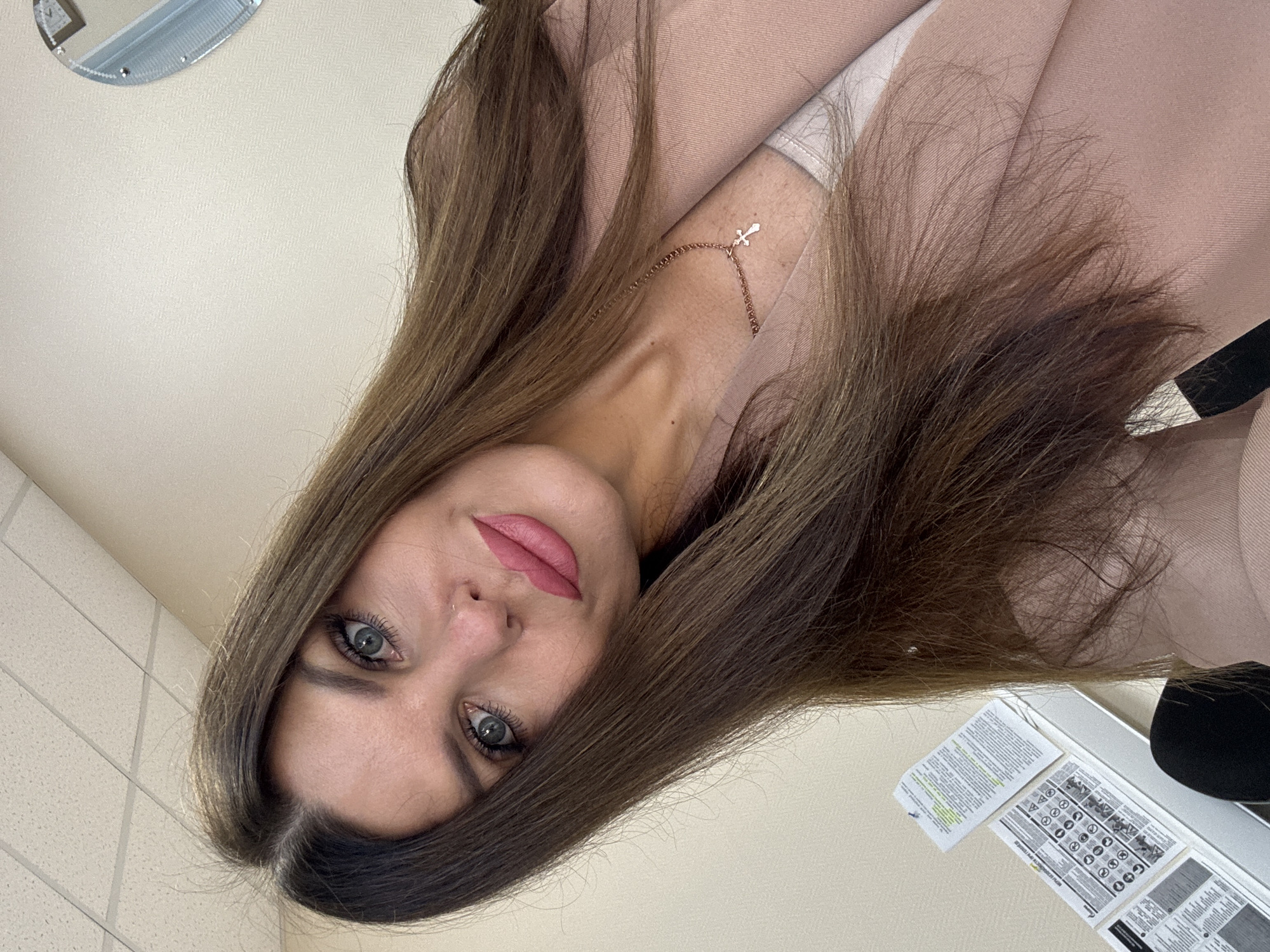 The image shows a woman with long, brown hair and blue eyes, wearing a pink blazer and a cross necklace. She is posing for a selfie, with her head tilted to the side. The background appears to be an indoor setting with a tiled ceiling and a plain wall. There are some papers on a surface to her left, and a mirror in the background. The scene is well-lit, suggesting it was taken during the day. The woman's facial expression is neutral, and her makeup is subtle. The overall impression is a candid, personal photograph. It is not possible to determine the specific location within the city of Chaikovsky, Russia, from the visual information provided.