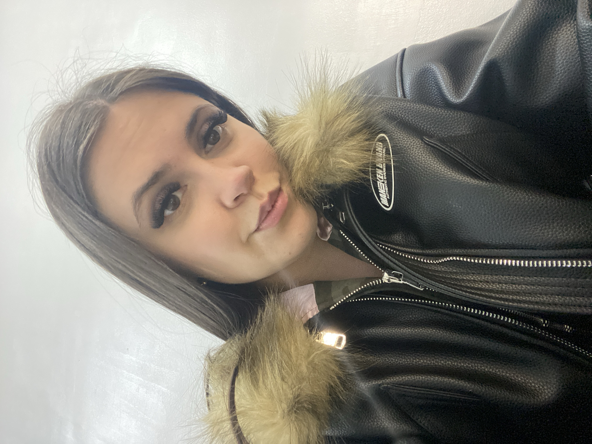 A young woman with dark hair and false eyelashes is wearing a black leather jacket with fur trim. The jacket has visible zippers and a patch. The woman is indoors, likely in Chaikovsky, Russia. The background is a plain white wall. The image is a selfie-style photograph.