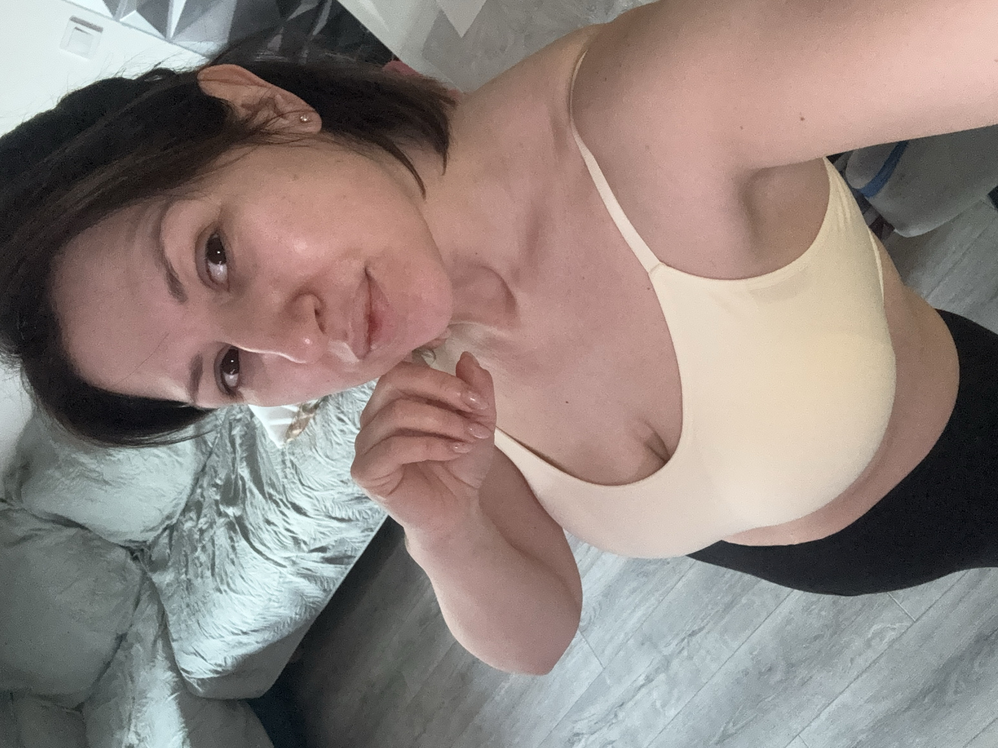 A woman with dark hair and fair skin is in a brightly lit interior space. She is wearing a light-colored sports bra and black leggings. Her right hand is near her face, and she is looking at the camera with a slight smile. The background includes a bed with a patterned gray bedspread and a light gray floor. The location is likely inside a residential building; there is no information to suggest the location is in the city of Sofia, Bulgaria.