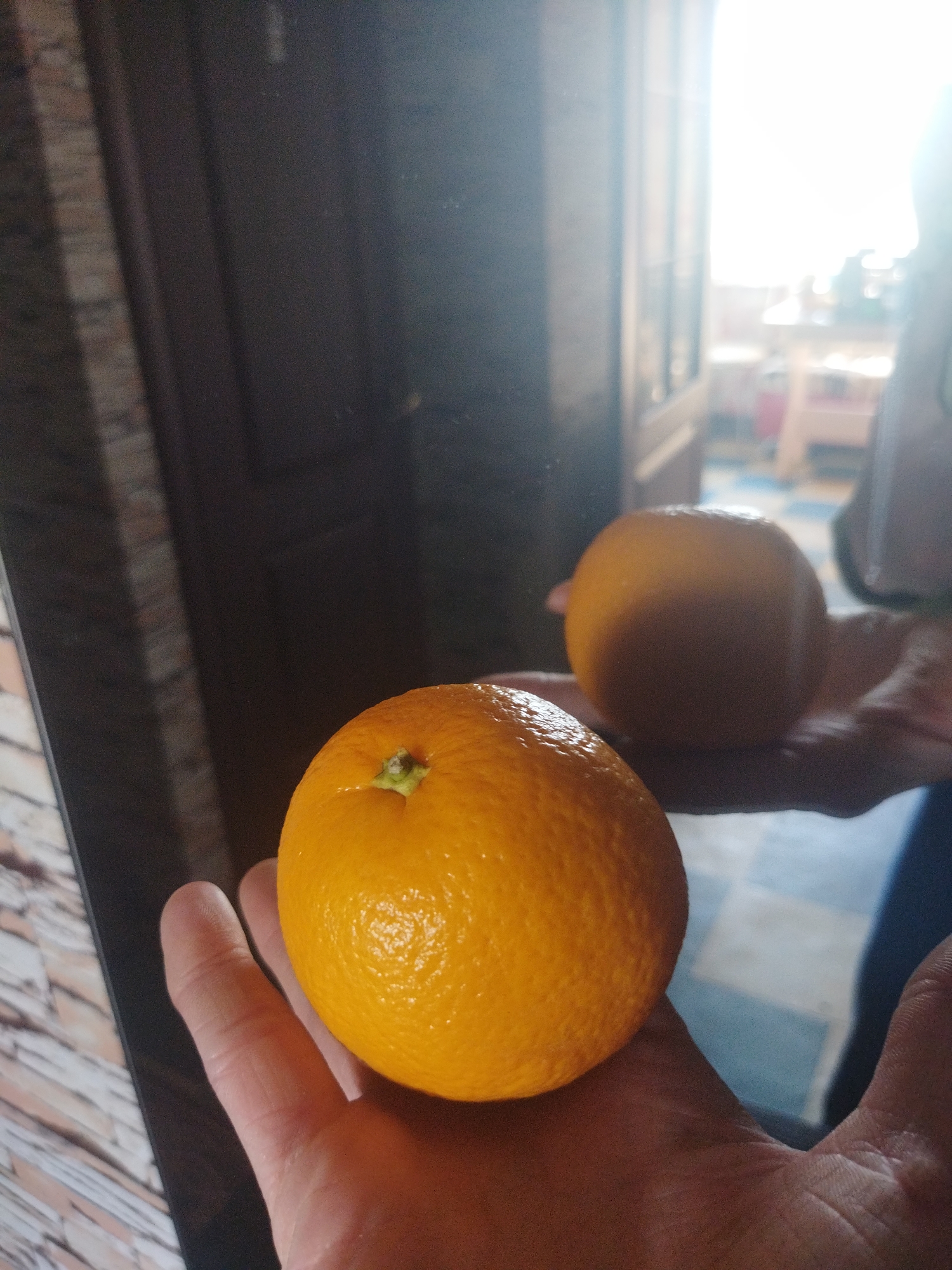 A hand holds an orange in front of a mirror. The reflection shows another orange and a blurry background, potentially a room interior. Kramatorsk, Ukraine is not discernible from the image.