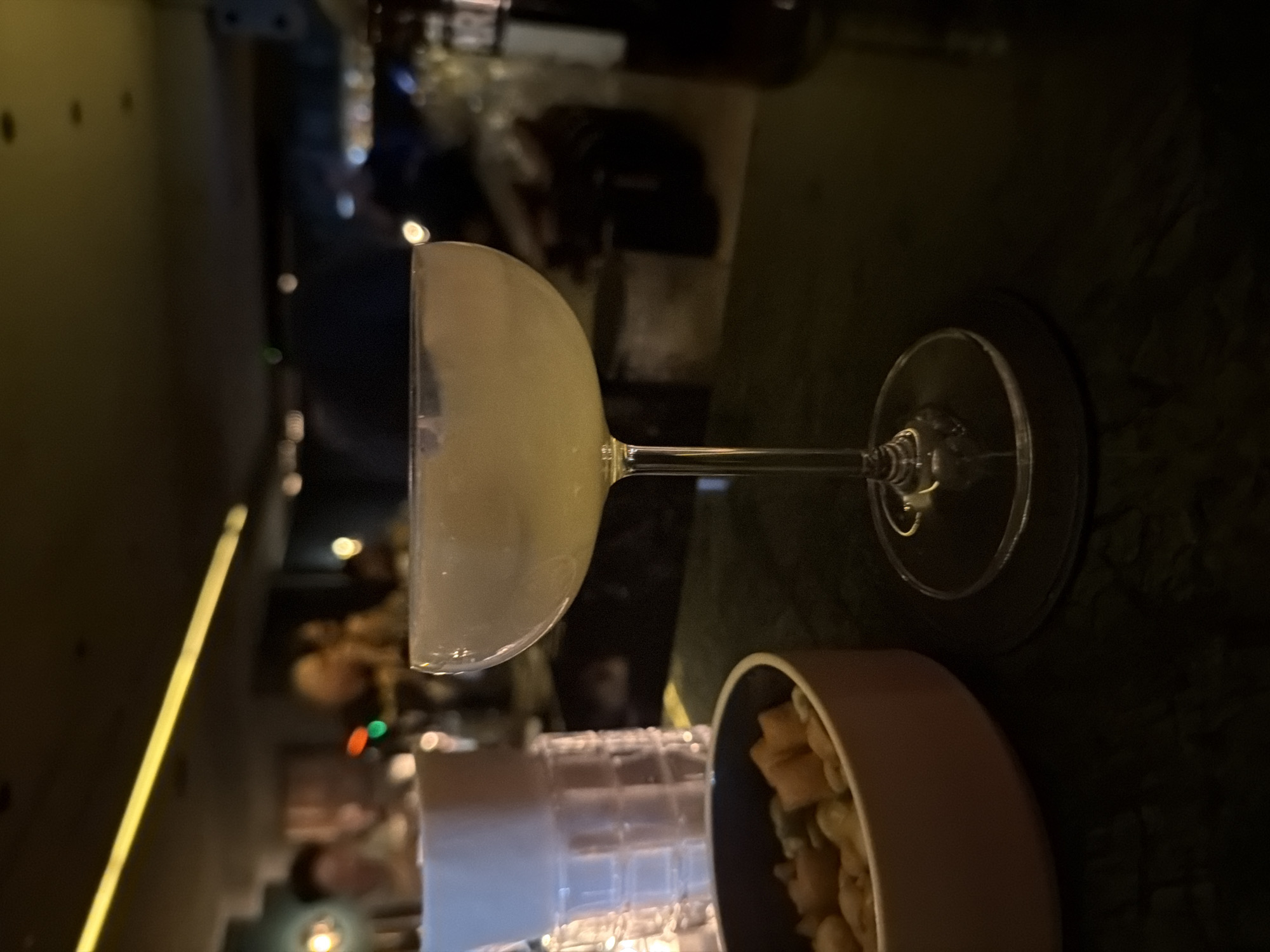 A cocktail glass sits nearly full on a dark surface, likely a countertop, accompanied by a small bowl containing what appears to be nuts or small snacks. The setting suggests a bar or restaurant, possibly in Bangkok, Thailand. The background is dimly lit, revealing blurred shapes hinting at other patrons and bar elements. A yellow light accentuates the left edge of the frame, adding depth.