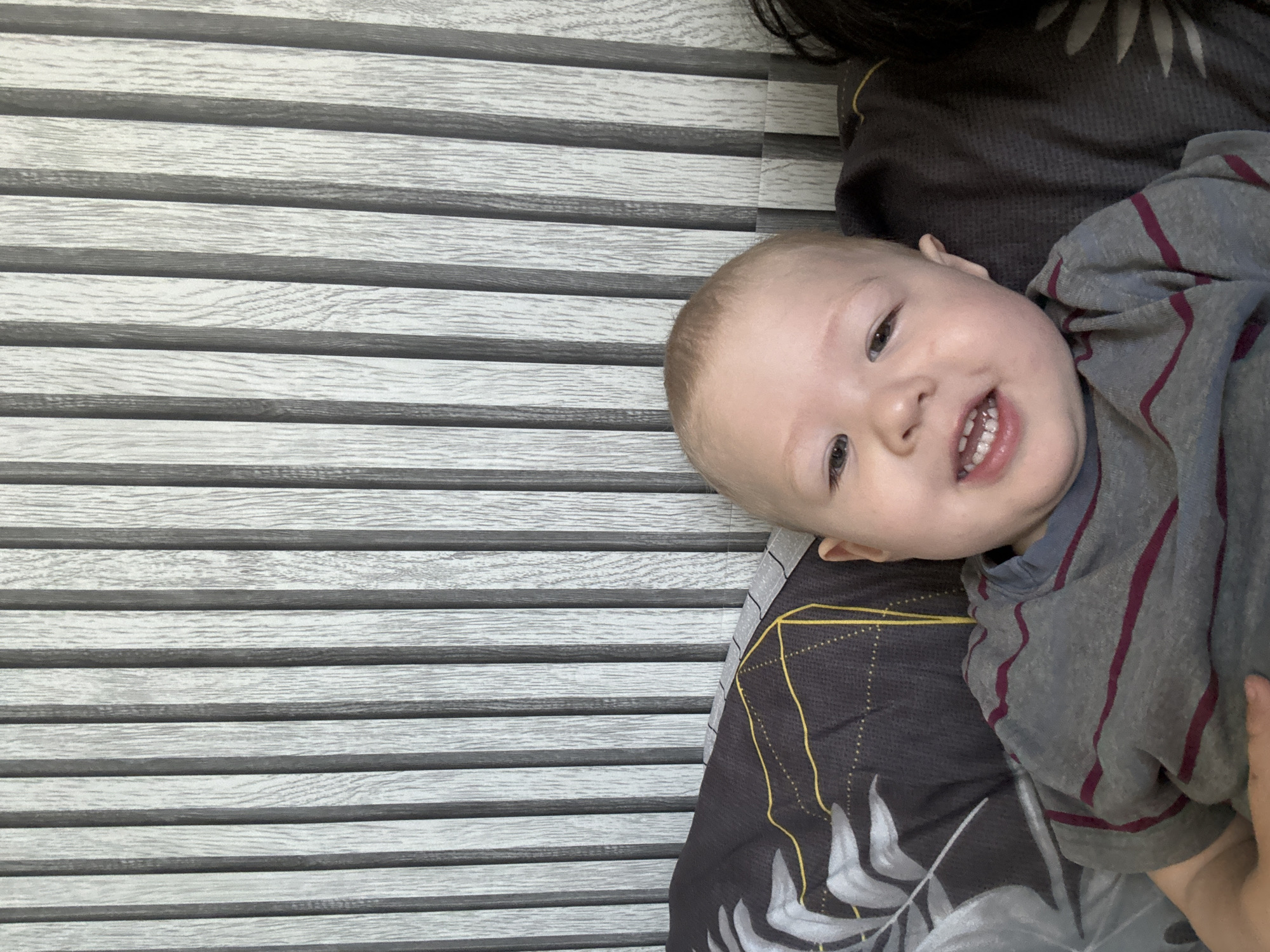 A close-up, high-angle, medium shot shows a fair-skinned baby boy with short, light brown hair and blue eyes, looking directly at the camera with a wide smile. He is wearing a gray t-shirt with thin, horizontal maroon stripes. The baby is positioned in the lower half of the frame, against a backdrop of a dark gray, textured wall with vertical, light gray wooden slats. To the left of the baby, a dark gray pillow with a black and white leaf pattern is visible. The overall impression is that of a casual, indoor photograph, likely taken in a residential setting. The specific location within Марковский, Russia, cannot be determined from the visual information provided.