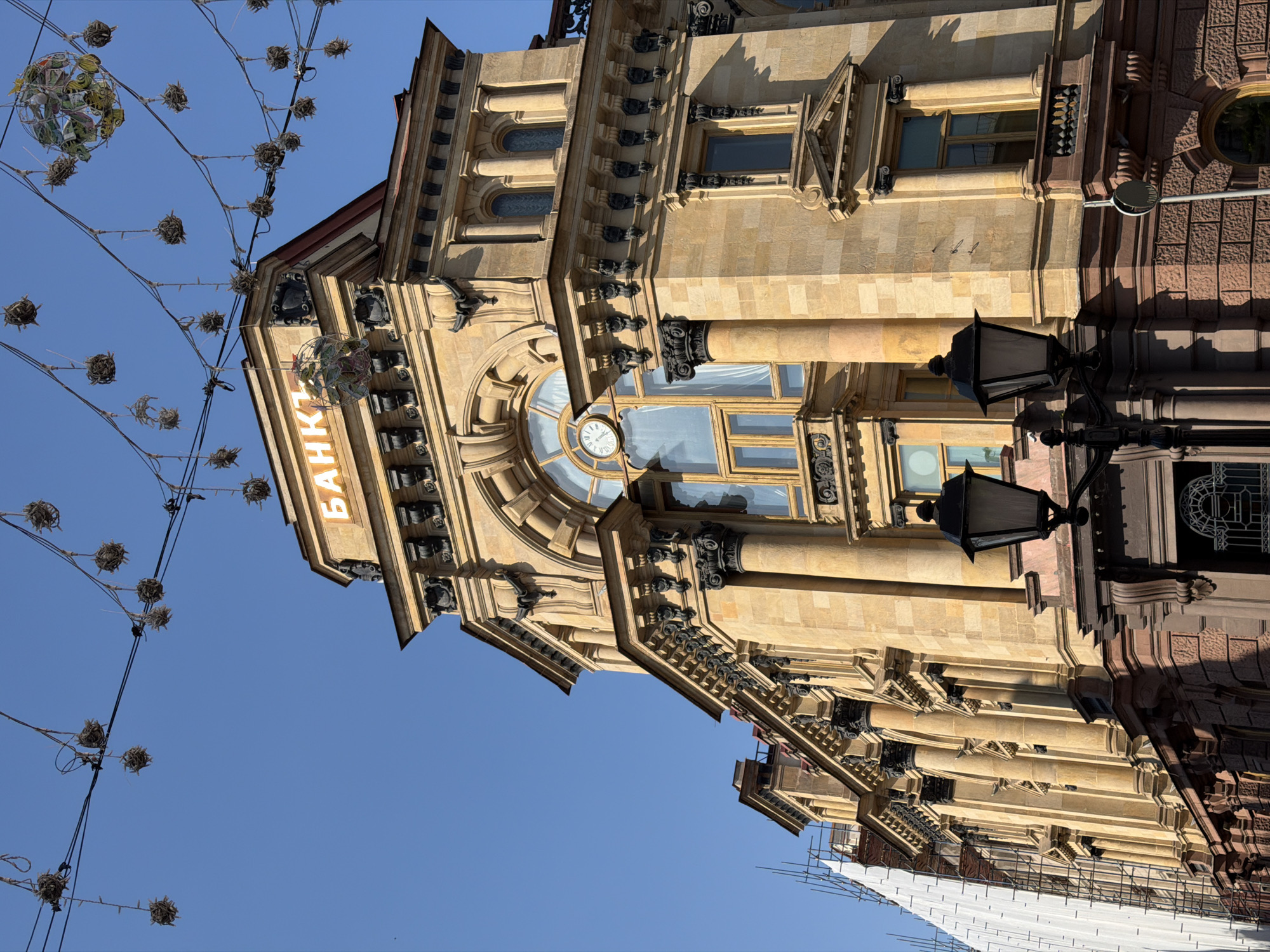 The image shows the Moscow Central Bank building in Moscow, Russia, as viewed from below. The building is made of tan stone, with brown stone on the right side. The structure has a clock in the center and the word "БАНК" on the left. There are lights hanging from wires above the building. The sky is blue. Scaffolding and white tarps are seen at the base of the building, suggesting ongoing construction. A black street lamp is in the foreground on the right.