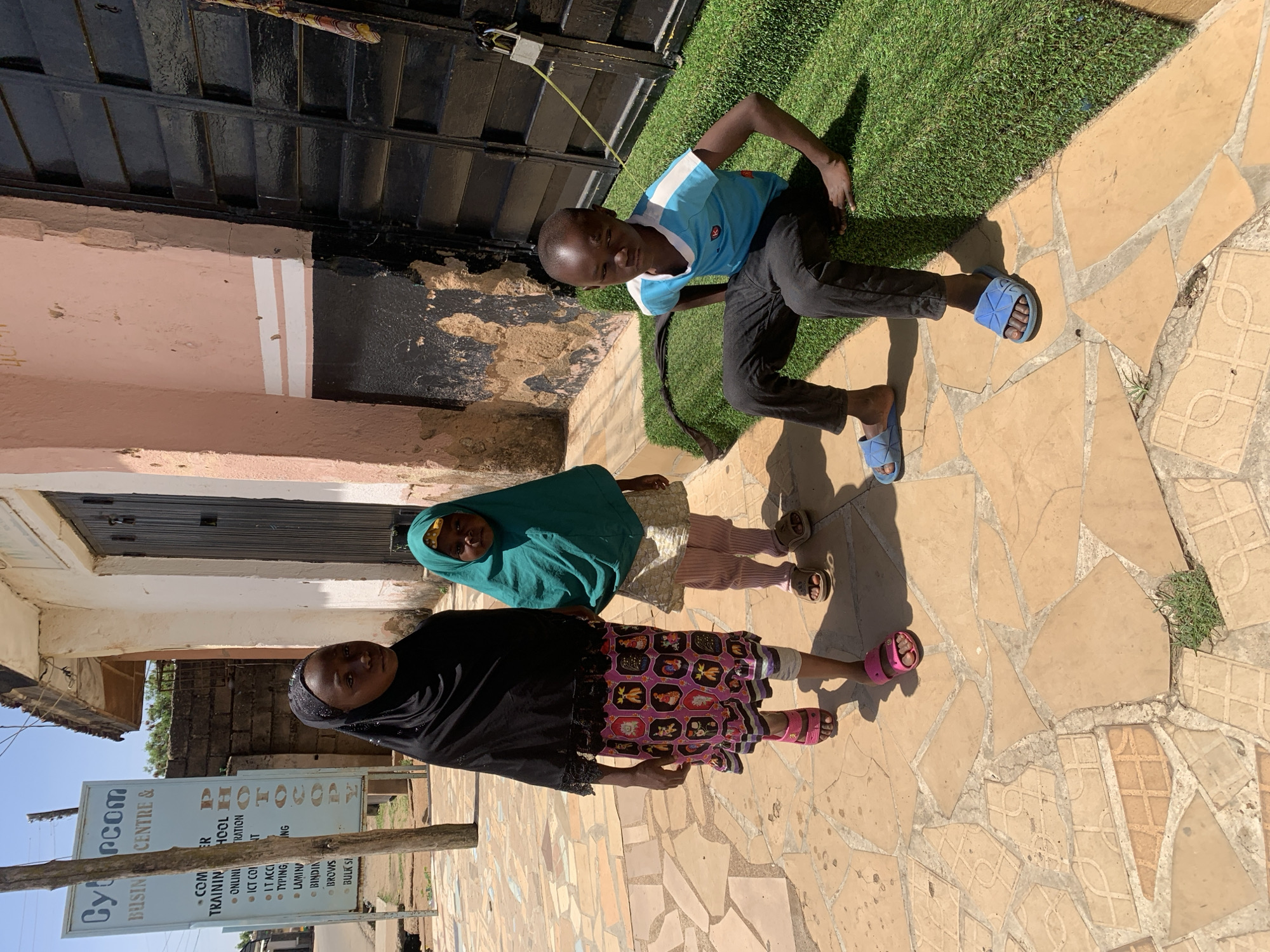 The image depicts three children standing in front of a building in Jos, Nigeria. Two girls are standing near the bottom of the frame; the girl at the bottom is wearing a black hijab and a skirt with colorful patterns. The girl next to her wears a green hijab. A boy is positioned near the top of the image, dressed in a blue shirt and black pants. He is wearing blue sandals. The ground is covered in paving stones. A sign for "Cybercom Business Centre & Computer Training School" is visible on the left side of the frame. There is also artificial turf on the upper right side of the image. A building with a black door is in the background.