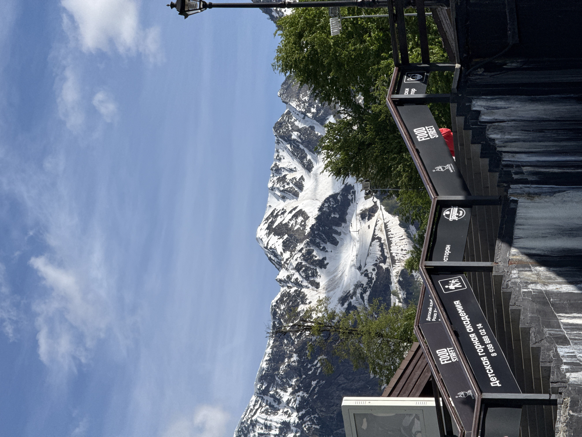 This vibrant daytime image captures a scenic mountain landscape combined with elements of a modern resort, likely in Estosadok, Russia. In the background, majestic snow-capped mountains dominate the view under a bright blue sky dotted with scattered white clouds. The mountains show distinct rocky textures partially covered by extensive snow and ice, suggesting either spring, early summer, or late autumn conditions.
In the mid-ground, lush green trees provide a contrast to the snowy peaks, hinting at warmer temperatures at lower elevations. The foreground features dark-colored architectural structures, including what appear to be steps or tiered platforms with railings, leading upwards to the right. A classic black lamppost with an ornate lantern is visible in the upper left corner, extending into the sky.
Several black signs with white text are prominent in the foreground and mid-ground, indicating various facilities. Readable text includes "FOOD STREET" repeated on multiple signs, suggesting a dining area. Other signs read "Детская горная академия" (Children's Mountain Academy) followed by a phone number "8 938 888 02 14," and "Детский клуб Роза Х" (Children's Club Rosa Khutor), identifying a specific resort area known for family activities. Partially visible text also includes "ресторан" (restaurant), "БАРБЕКЮ" (BBQ), and "Груши." One sign features an icon of a person with outstretched arms, possibly indicating an activity or welcome.
A glimpse of a person wearing a red jacket can be seen behind one of the signs and railings, adding a touch of human presence to the otherwise natural and architectural scene. In the lower left, a portion of a white object, possibly a monitor or display screen, is visible. The bright lighting and clear sky suggest pleasant weather, ideal for outdoor activities in this mountain resort setting.