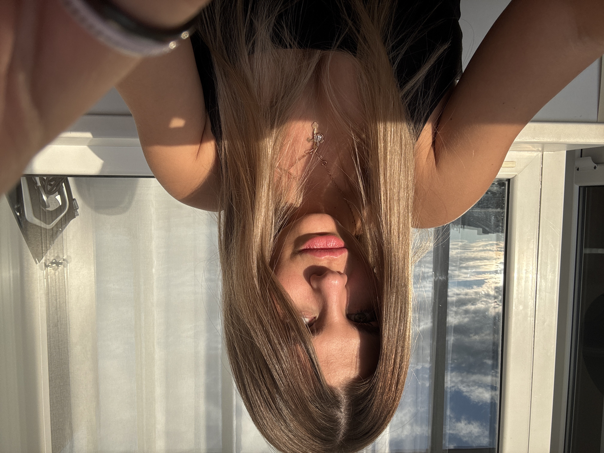 A young adult female with long blonde hair and fair skin is captured in a selfie, positioned centrally in the frame. She is wearing a black tank top and a delicate necklace with a cross pendant. The lighting suggests it is daytime, with sunlight casting shadows across her face and arms. Behind her, a large window reveals a cloudy sky and urban landscape, indicating an indoor setting, possibly an apartment or hotel room, with a view of Sarapul, Russia. The overall mood is casual and personal.