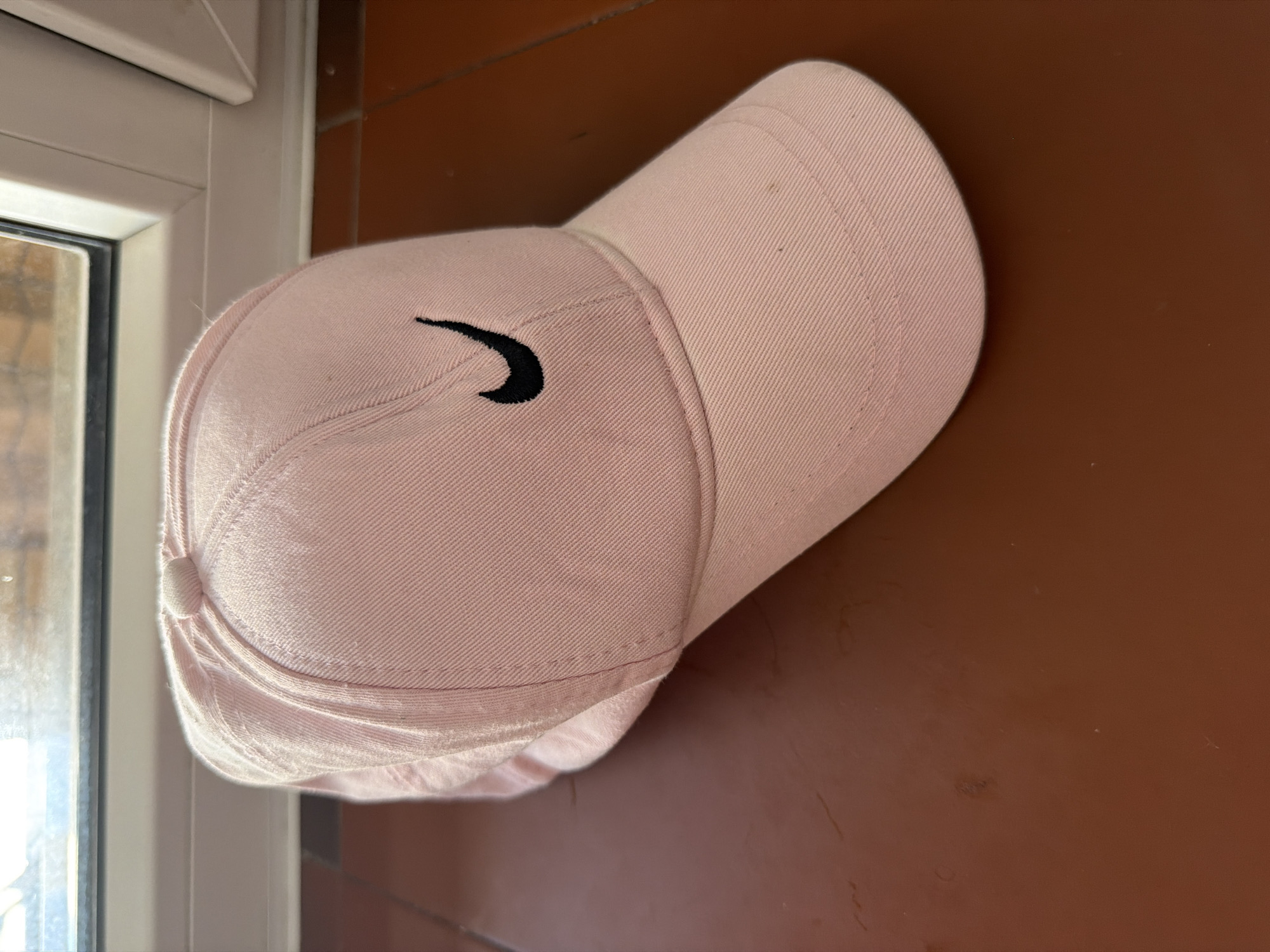 A close-up, slightly high-angle shot shows a pink Nike baseball cap with a black swoosh logo placed on a reddish-brown, smooth surface. The cap is angled, with the brim facing towards the viewer. The background features a window frame and what appears to be an outdoor scene with muted colors, possibly a window in Qarağandı city administration, Kazakhstan. The lighting suggests an indoor setting with natural light coming from the window. The surface beneath the cap has a few smudges and light-colored specks.