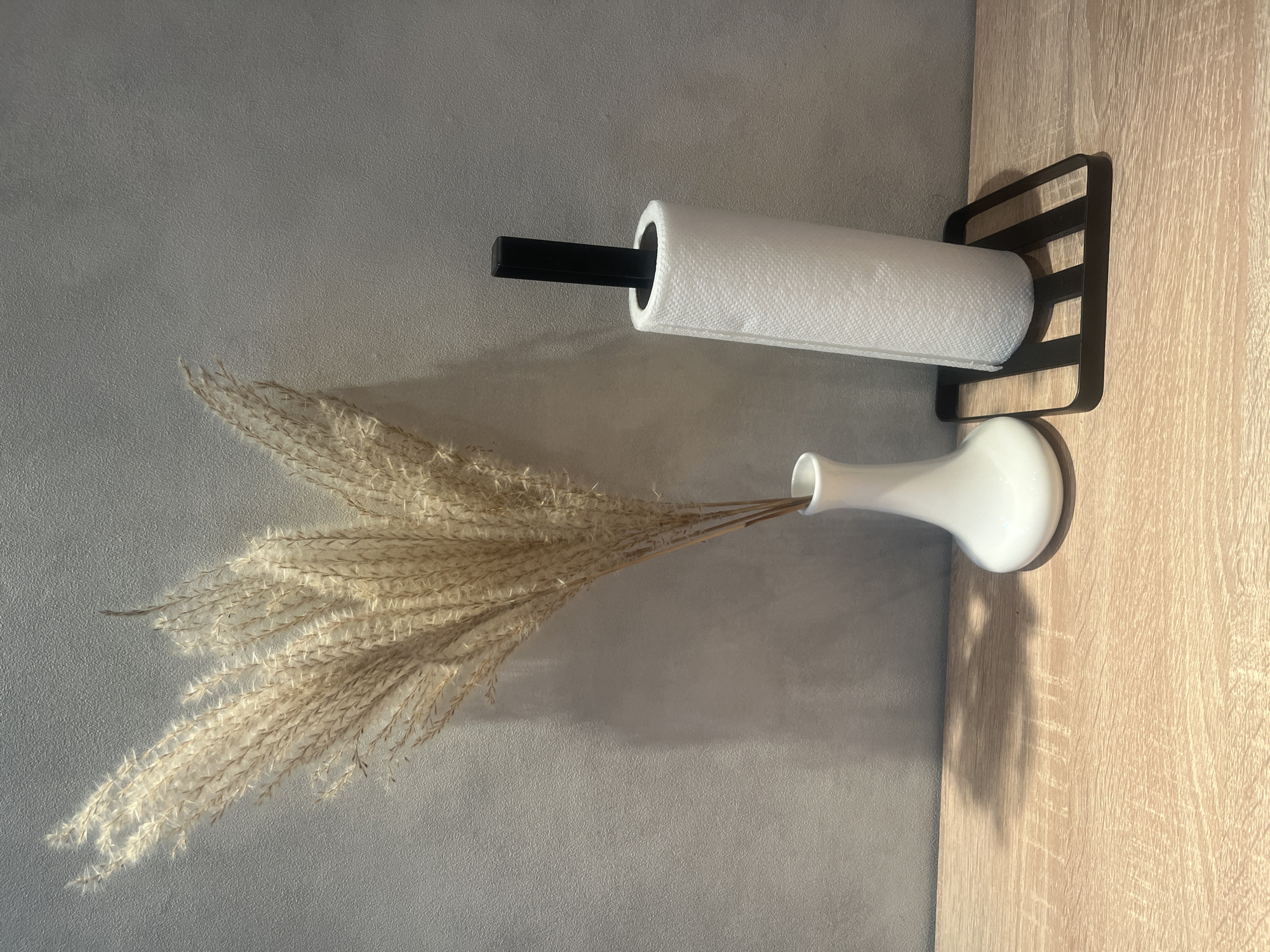 The image shows a still life composition on a wooden countertop against a textured gray wall. On the left, a white ceramic vase holds a large, dried pampas grass plume. To the right of the vase is a black paper towel holder with a roll of white paper towels. The lighting casts shadows on the wall and countertop, suggesting an indoor setting, possibly a kitchen or living area. The scene is static and shows no human presence. The location in Rybnitsa, Moldova is not evident from the visual information presented in the image.