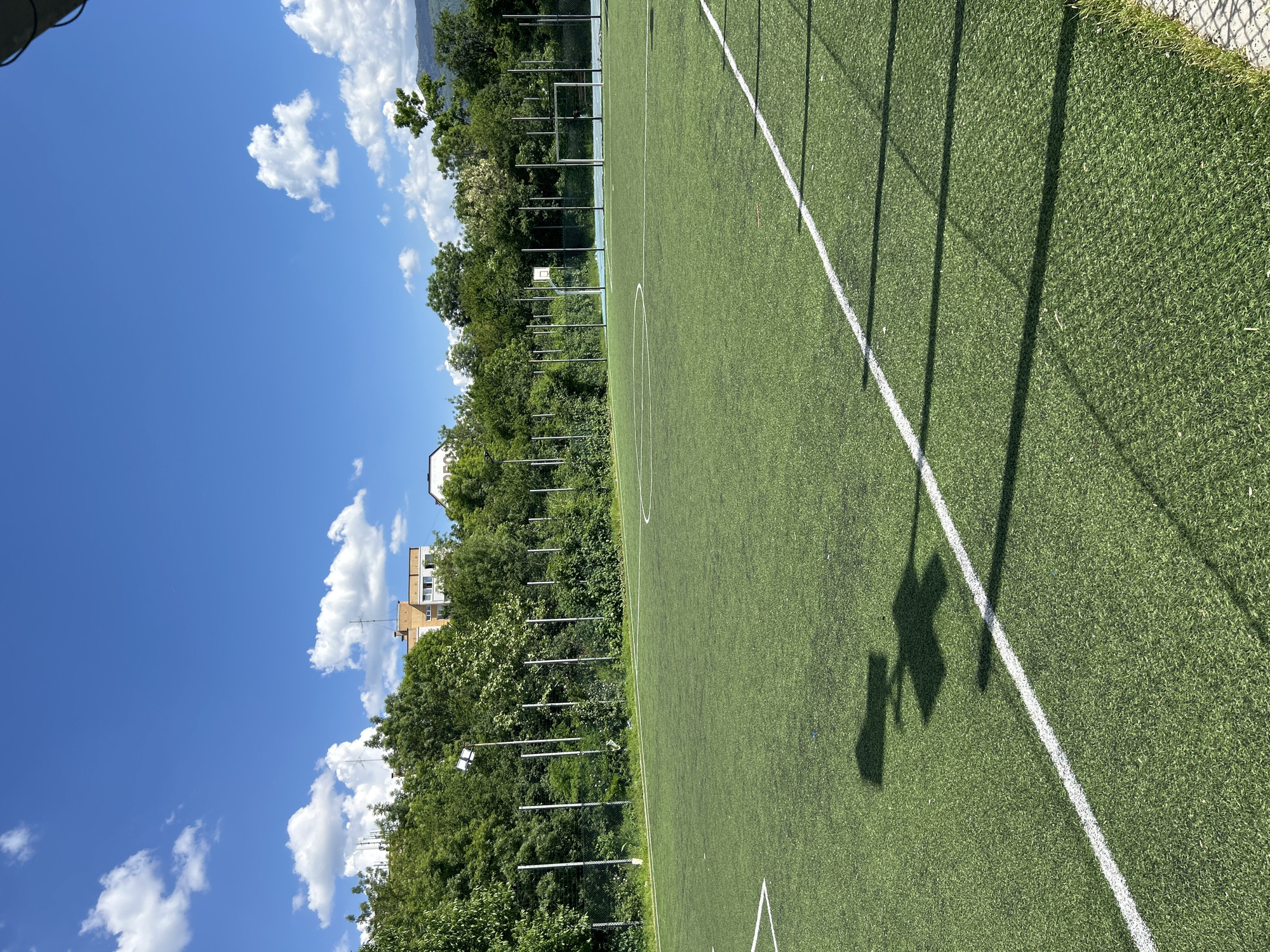 The image shows a synthetic turf sports field in an unidentified city. The field is marked with white lines and a center circle. A fence runs along the right edge of the field, and the shadows of the fence posts are cast onto the turf. Beyond the fence and field are trees, a building, and a blue sky with clouds.
