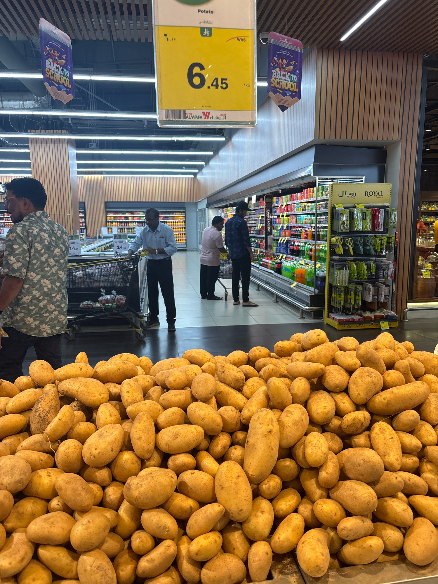 The image depicts the produce section of a supermarket in Dammam, Saudi Arabia, likely Al Othaim Market, given the signage. In the foreground, a large pile of fresh, unwashed potatoes is displayed. Behind the potatoes, several shoppers are visible. A man in a floral shirt stands near a shopping cart filled with goods, looking towards the right. To his right, a man in a blue shirt pushes a shopping cart, interacting with the products on a shelf. Further in the background, other shoppers are browsing the aisles, which are stocked with various food and drink items. Overhead, a "Back to School" promotional banner hangs, shaped like a pencil. The store environment appears well-lit and organized.