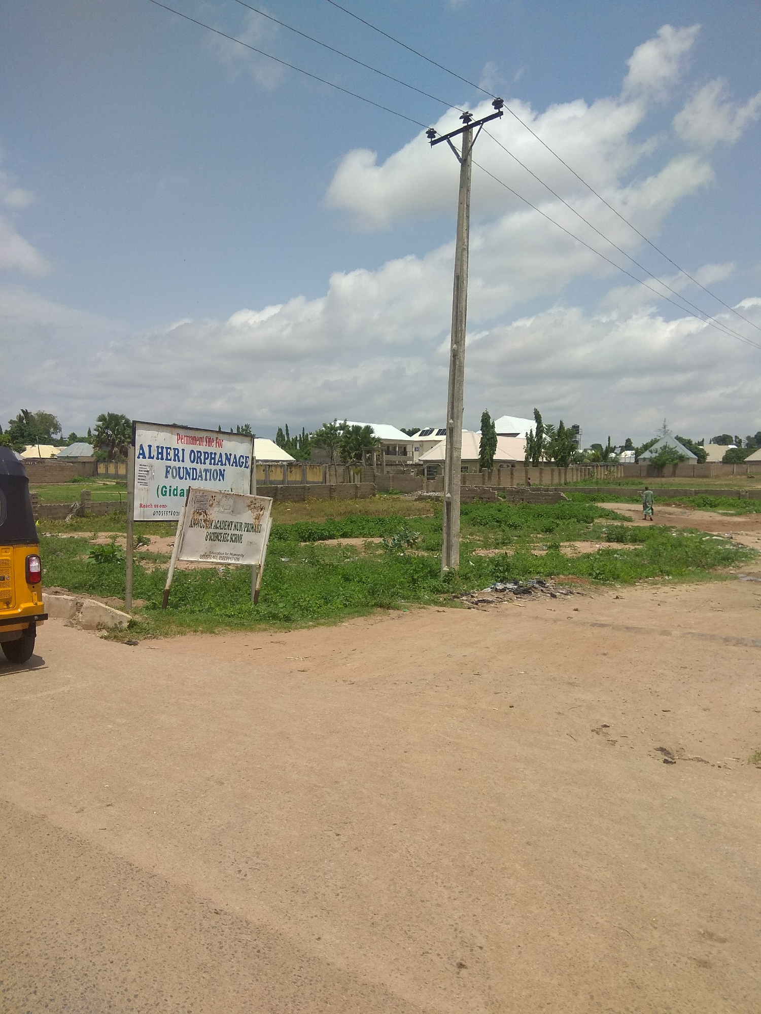 In Jalingo, Nigeria, a yellow motorized tricycle is parked on the left side of a dirt road. Two signs are erected in front of a grassy field. The larger sign reads "Permanent Site For ALHERI ORPHANAGE FOUNDATION (Gida)". The smaller sign, beneath it, states "ALHERI ACADEMY NUR. PRIMARY & SCIENCE SEC. SCHOOL". Behind the field, residential buildings with white roofs and some under construction are visible. A utility pole with power lines stretches across the frame against a partly cloudy sky. A man in a green garment walks in the distance on the right side of the scene.