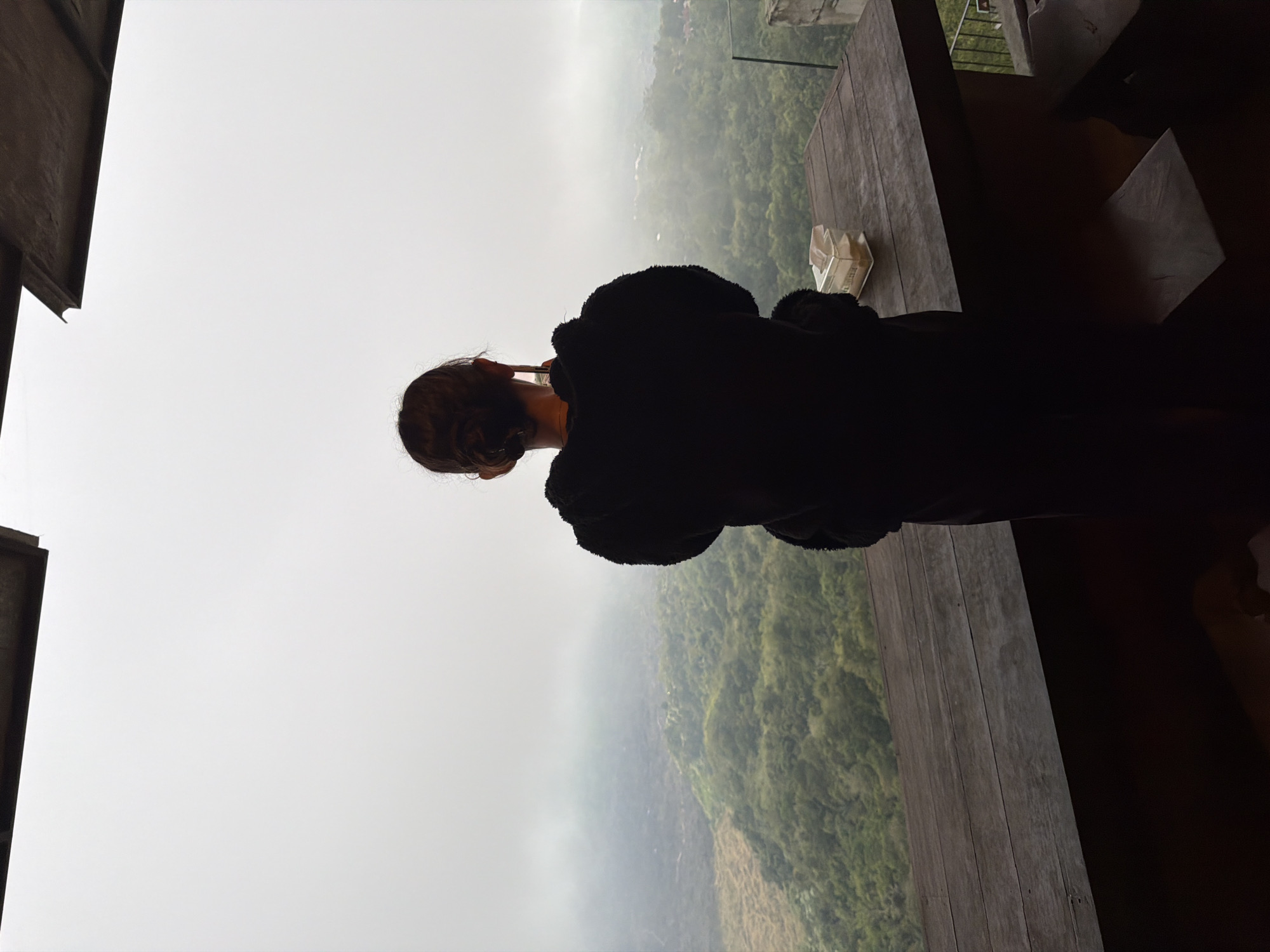 A person stands on a wooden deck, facing away from the camera, looking out at a foggy mountainous landscape. The person is wearing a black, fuzzy hooded jacket. On the wooden table in front of them is a clear plastic container filled with yellow objects, possibly food or tissues, and a white folded napkin. The background shows lush green trees and rolling hills obscured by a thick layer of fog. The sky is overcast and white. A portion of a dark, angled structure is visible at the top of the frame. This scene appears to be from a viewpoint in Kintamani, Indonesia, known for its scenic views of mountains and lakes.