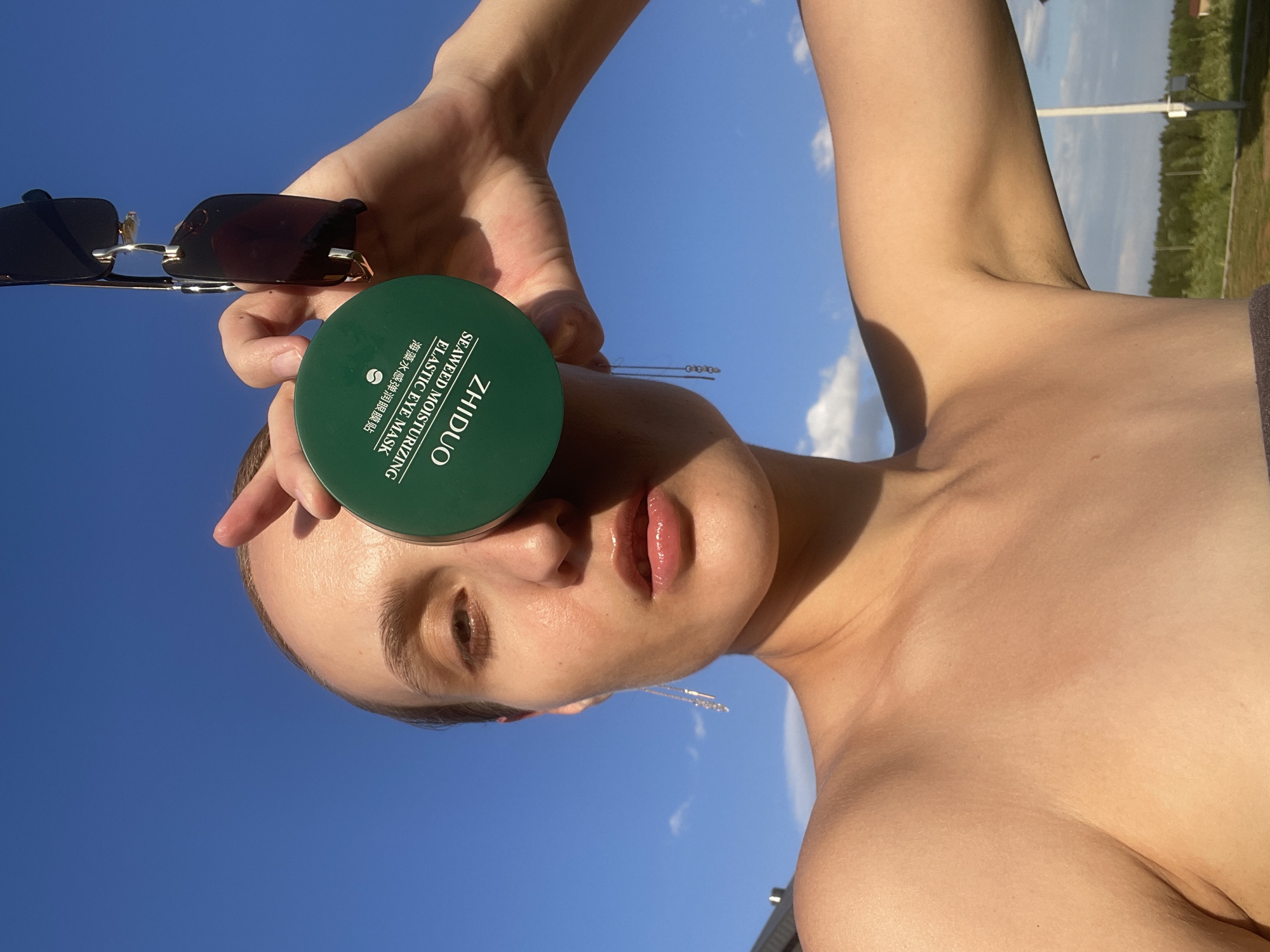 The image shows a fair-skinned individual holding a green jar of "ZHIDUO Seaweed Moisturizing Elastic Eye Mask" over one eye. They also hold a pair of dark sunglasses in the same hand. The person's face, chest, and arm are visible, illuminated by strong sunlight. The sky is clear and blue. In the background, there are trees and a grassy area. Given the prompt's specification, the location is Подшивалово, Russia.