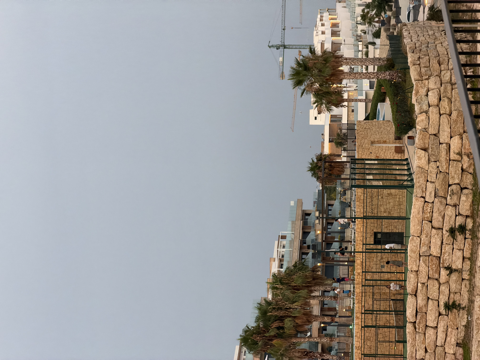 The image depicts a resort or residential complex under construction in Finestrat, Spain. The foreground features a low, dry-stone wall constructed from large, rough-hewn beige stones. Behind the wall, a paddle tennis court is visible, enclosed by green fencing. Several individuals are present on and around the court, with some appearing to be playing paddle tennis and others standing on a raised walkway.
In the mid-ground, several modern buildings with white facades and glass balconies are visible, interspersed with palm trees. Construction cranes are prominent in the background, indicating ongoing development in the area. The sky is overcast and a pale gray. The overall impression is one of a coastal development in an early evening light.