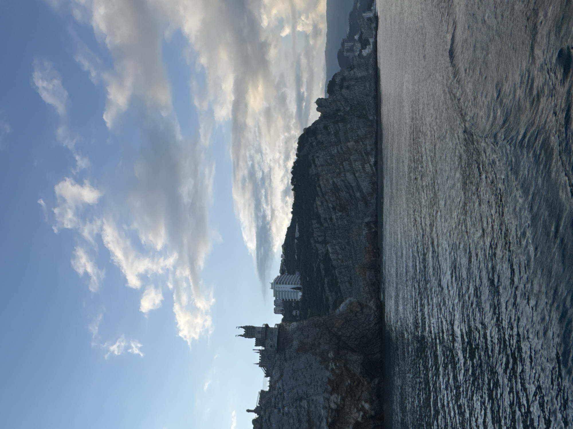 The image depicts a coastal scene with a prominent rocky cliff on which a castle-like structure is perched. To the left is the blue sky with white clouds. The right part is filled with the surface of the sea. The location is Гаспринский поселковый совет, Ukraine.