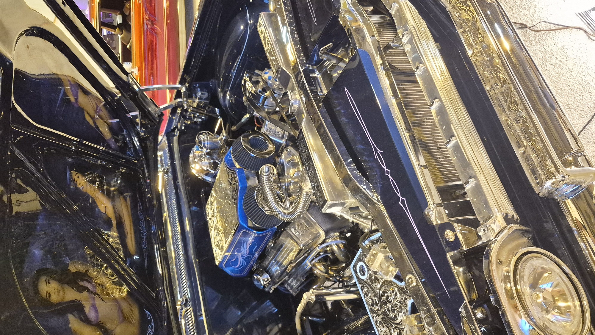 The image displays the engine bay of a custom black car, likely a classic American model, with extensive chrome and intricate engraved detailing. The hood is open, revealing a highly polished engine with a blue air intake system adorned with gold engraving. The car's interior, visible through the windshield, features pinstriping and images of scantily clad women painted on the inside of the hood and dashboard. The background shows other vehicles, including a red car, suggesting this is an automotive event or exhibition. The lighting is artificial, creating reflections on the polished surfaces. The scene is devoid of any specific landmarks that would indicate its location within Sacramento, California, USA.