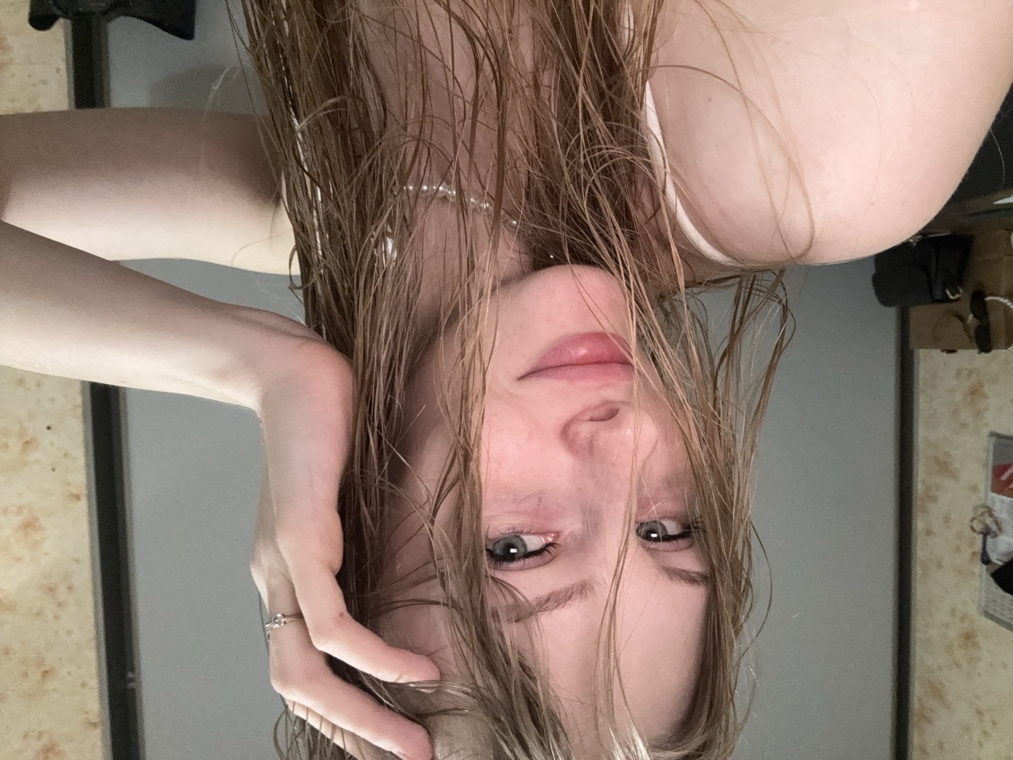 A fair-skinned young adult female with long, wet, light brown hair is centered in a close-up, self-portrait image. Her hair is disheveled, with individual strands clinging to her forehead and shoulders, indicative of recent exposure to water. Her blue-grey eyes are directed straight at the camera, with a slight redness noticeable beneath the lower lash line. She exhibits a subtle, closed-mouth smile. A small, simple metallic ring is present on the ring finger of her right hand, which is positioned with fingers splayed, appearing to run through or push back her wet hair from her forehead. A delicate, white beaded or pearl-like necklace encircles her neck. She wears a white, thin-strapped garment, possibly a camisole or tank top, with her left shoulder and upper arm partially visible in the foreground.
The subject is situated indoors. The background consists of two distinct wall sections: to the left, a light-colored, possibly textured or patterned wallpaper in muted beige/brown tones, with blurry, dark shapes that suggest hanging objects or furniture; to the right and directly behind the subject, a plain, smooth, light grey wall. A vertical dark line on the far right may indicate a door frame or the edge of a mirror. In the blurry far left background, several items are discernible on a surface, including a light brown paper bag and a dark-colored shoulder bag or clutch, with what appears to be sunglasses resting atop the brown bag. The lighting is soft and even, suggesting an indoor artificial light source. The setting is identified as being in Perm, Russia.