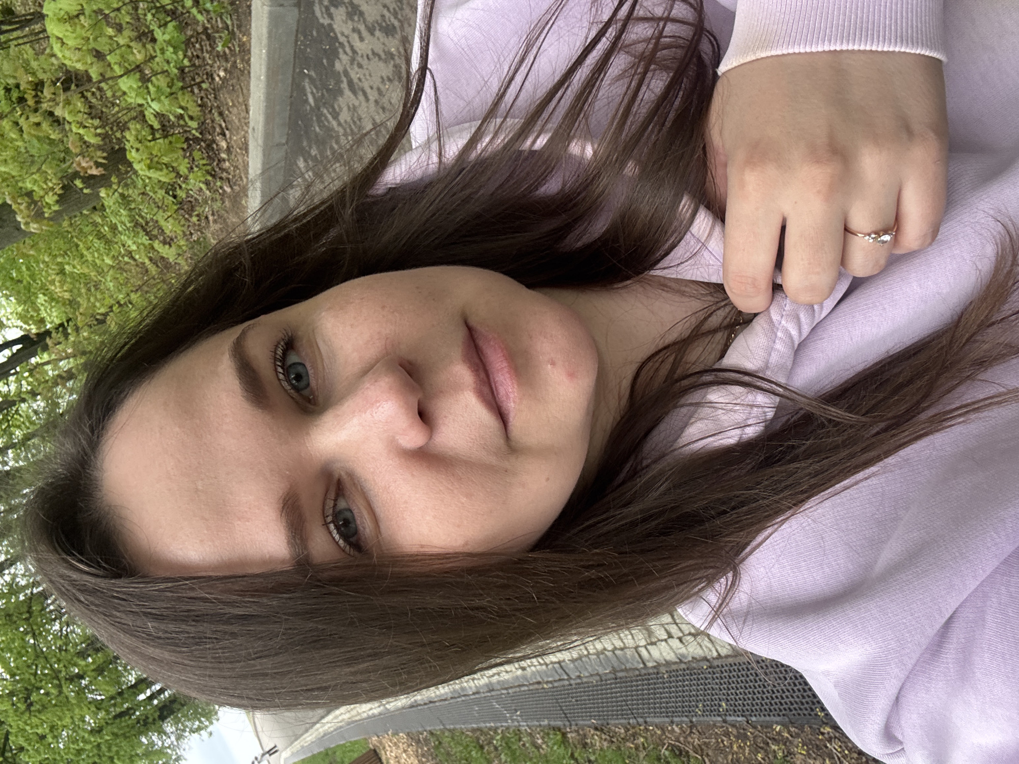 A close-up, slightly canted, selfie of a woman with brown hair and light eyes. She is wearing a light purple hooded sweatshirt. On her right hand is a delicate ring with what appears to be three small stones. The background shows greenery, possibly trees and grass, along with a paved area. Based on the available details, it is impossible to confirm the location in Nizhny Novgorod, Russia.