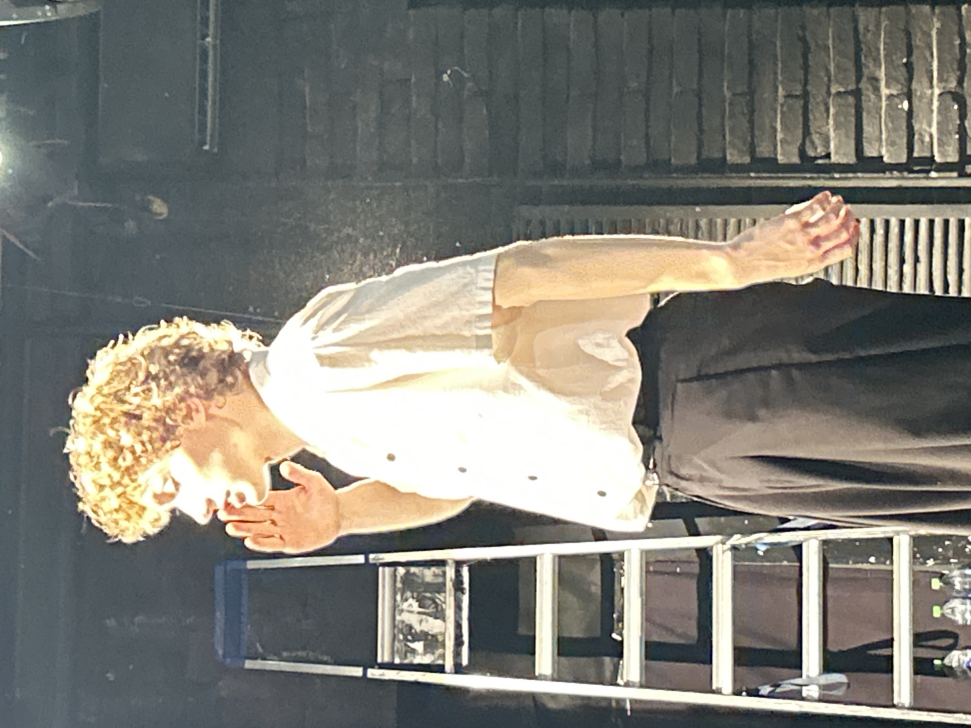 A young man with curly, blonde hair stands in profile, facing left, with his right hand cupped around his mouth, as if speaking or calling out. He is wearing a loose, white, short-sleeved shirt buttoned down the front and dark, wide-legged trousers. His left arm hangs loosely at his side. Behind him, to his left, is a silver stepladder with blue supports. The background appears to be a dark stage set with brickwork on the right. The lighting is dramatic, with a spotlight illuminating the man from the front and left. The image is a medium shot, focusing on the man from the chest up.