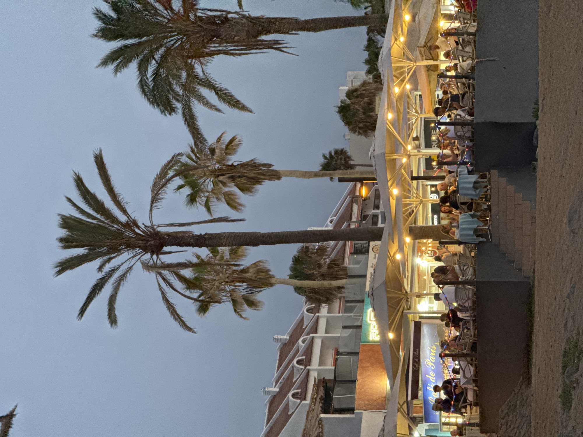 The image depicts an outdoor seating area of a restaurant or cafe at dusk in Arona, Spain. Several large umbrellas, illuminated by string lights, provide cover for tables where patrons are seated. Palm trees are visible in the background, with a building complex behind them. People are seen dining, socializing, and being served. The overall atmosphere suggests a relaxed evening ambiance.