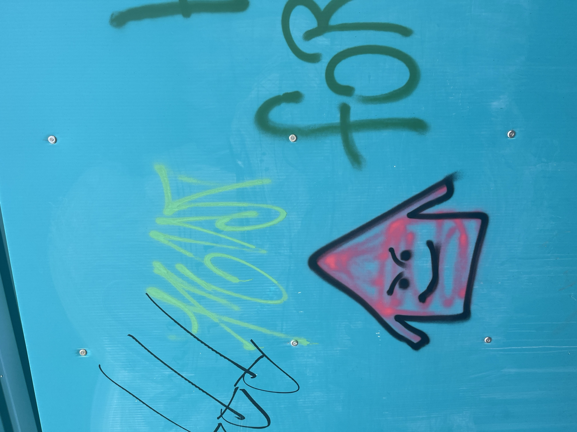 The image is a close-up shot of a teal-colored corrugated metal surface adorned with graffiti. On the left, there are black abstract scribbles. To the right of these, in light green spray paint, are stylized characters that are difficult to decipher. Below the green graffiti, a cartoonish, house-shaped figure with a pink and red interior and a black outline is spray-painted. This figure has angry eyes and a wide, smiling mouth. Further to the right, in dark green spray paint, are the letters "for". The metal surface has visible screws or rivets at various points. The lighting suggests it is daytime. The overall impression is that of an urban environment, possibly a wall or a structure in a public space. There are no people visible in the image. The graffiti appears to be recent.
