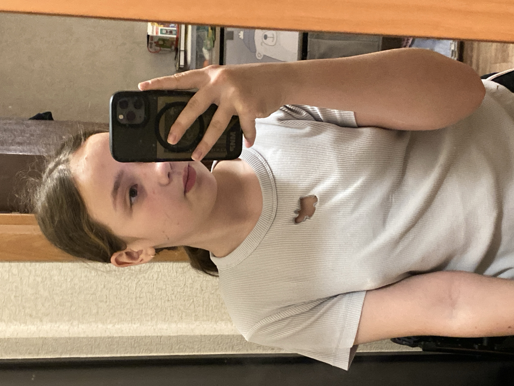A person with light brown hair and light skin is holding a smartphone in their right hand and taking a selfie in front of a mirror. The person is wearing a white t-shirt with a small bear cut-out near the neckline. The background shows a wall with a textured pattern and a shelf with various items. The mirror frame is wooden. The location is Unknown, Unknown.