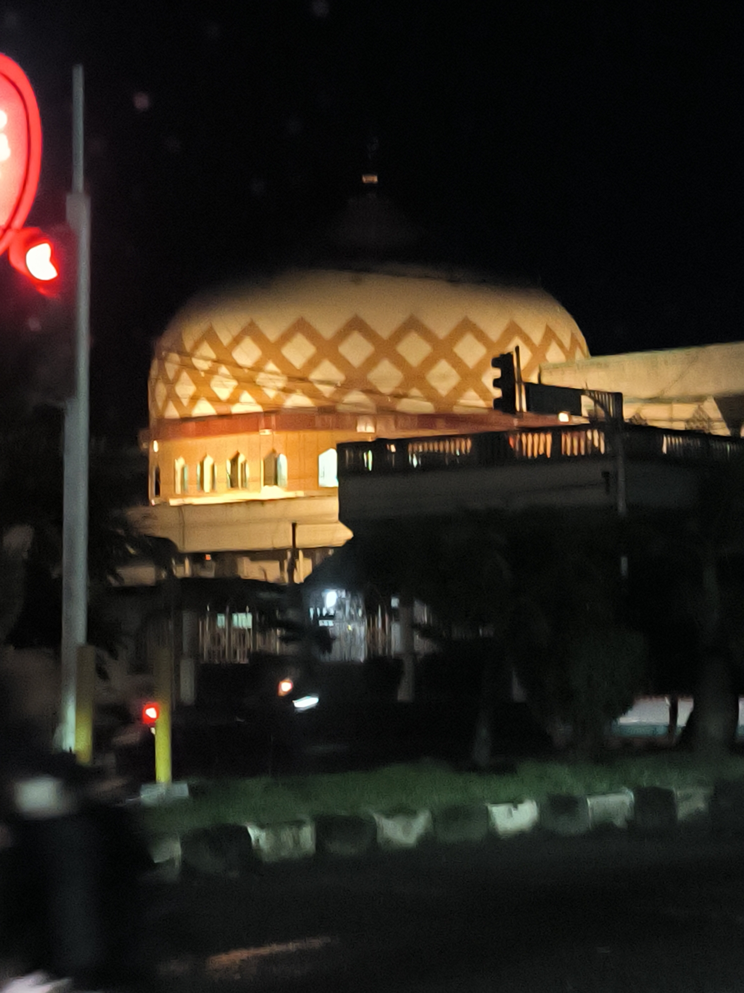 The image captures a nighttime view of what appears to be the Islamic Center Mosque in Lhokseumawe, Indonesia. The mosque's distinctive dome with a geometric pattern is illuminated. The image is somewhat obscured by foreground elements, including trees and a portion of a traffic light showing red. The scene suggests an urban environment with the mosque as a prominent structure. A row of low concrete barriers is visible in the foreground. The image is generally dark due to the nighttime setting.