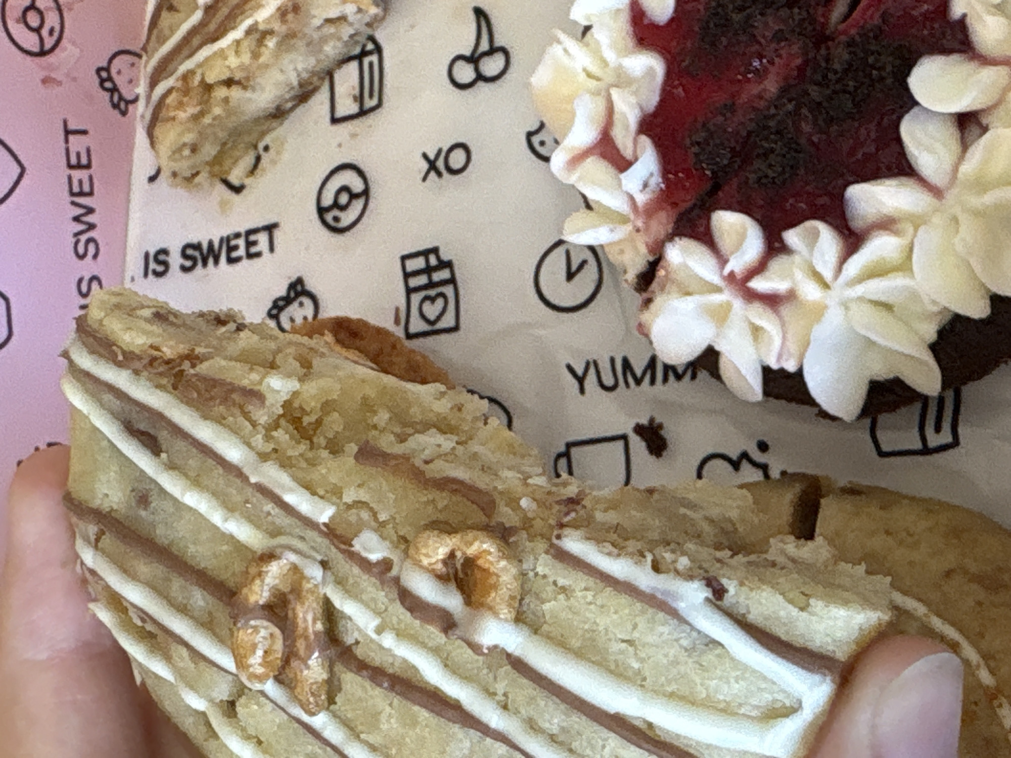 This media file features a close-up view of several pastries, likely cookies or donuts, against a patterned background. The primary subjects are the baked goods themselves, showcasing their textures and decorative elements. One pastry in the foreground is a light-colored cookie, bisected, with white and brown stripes drizzled across its surface. Small, nut-like pieces are embedded in the cookie. To the right, a darker, possibly chocolate, pastry is visible, topped with a rich red filling and adorned with piped white frosting. The background is a white surface with a repeating pattern of small black icons. These icons include stylized cherries, a coffee cup, a clock, a chocolate bar with a heart, and outlines of donuts. Text is also visible on the background. On the left, in a pink area, the words "IS SWEET" are printed vertically, and further to the right, "YUMM" is printed horizontally. A hand is visible at the bottom left, holding one of the pastries. The lighting suggests it is daytime, and the overall impression is of a sweet treat being presented or consumed. The provided location context of Saint Petersburg, Russia, does not offer any specific visual cues within the image itself. The emotions conveyed are likely those associated with enjoying dessert – pleasure and satisfaction.