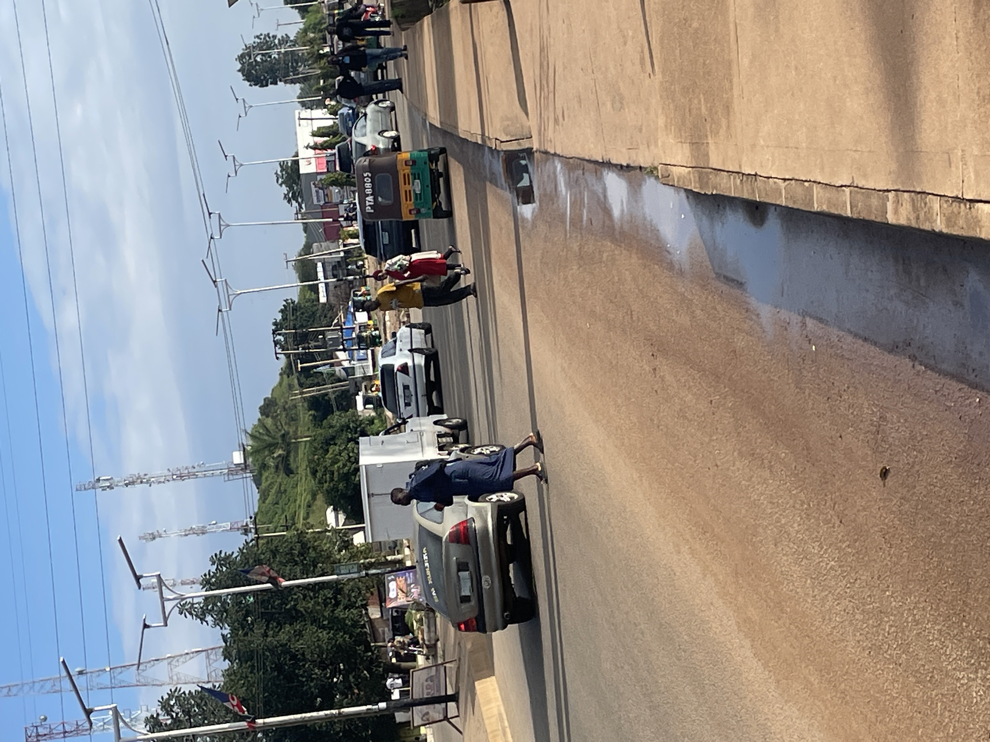 The image depicts a street scene in Jos, Nigeria, on a clear day with a partly cloudy sky. The foreground shows a dusty road with a concrete gutter on the right side, containing some accumulated water and debris. A person in a dark blue robe and headscarf is crossing the road, with a backpack on their shoulders, moving from right to left. Behind them, a silver sedan with tinted windows is parked, and a white truck with a covered bed is also visible.
Further down the road, several vehicles are present, including a green and yellow auto-rickshaw with the license plate "PTA 0805," a dark SUV, and other cars. A group of people, including a man in a yellow t-shirt and a woman in a red dress, are walking on the sidewalk. In the background, there are numerous cell towers and utility poles with electrical wires stretching across the frame. Trees and buildings are scattered throughout the landscape, with a green, hilly area in the far background. The overall impression is a busy urban environment.