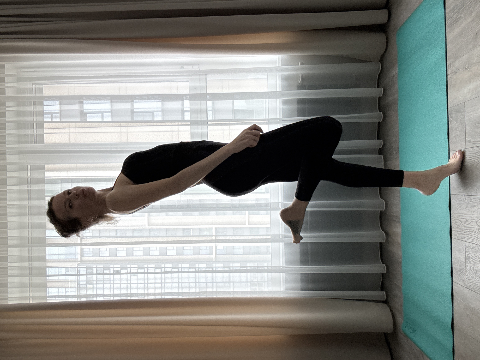 A woman is standing on a wooden floor in a room. She is wearing a black leotard and black leggings. Her left leg is bent at the knee and her foot is resting on her right calf. Her right leg is extended straight out behind her. She is facing the camera with her head tilted to the left. Her hair is pulled back in a bun. She has a tattoo on her left foot. A turquoise yoga mat is on the floor in front of her. The background of the image consists of a large window covered by sheer white curtains and beige curtains on the sides. Outside the window, buildings are visible.