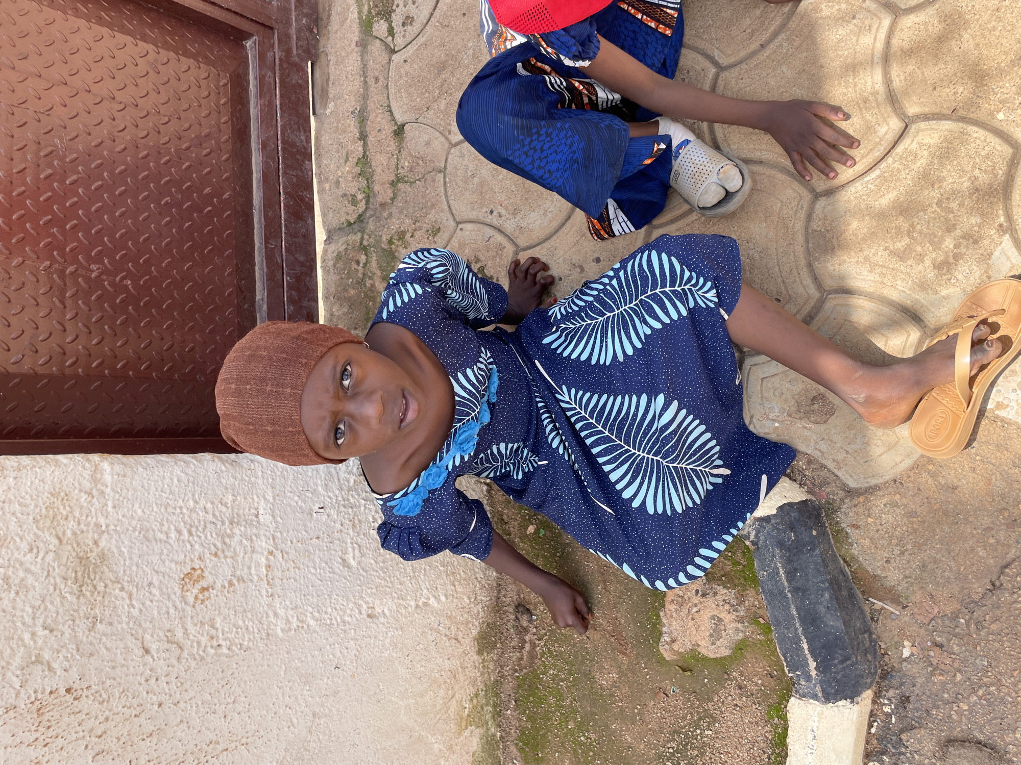 The image shows two young girls sitting on patterned paving stones in front of a textured, light-colored wall and a dark red metal door. The girl in the foreground, wearing a brown knit cap and a dark blue dress with light blue leaf patterns, is looking towards the camera. Her right arm is resting on the ground, and her left hand is on her leg. She is wearing tan flip-flops. To her right, another girl is partially visible, wearing a similar blue dress and white socks with grey sandals. Her hands are on the paving stones. The environment appears to be outdoors in a sunny location, likely in the city of Jos, Nigeria, given the common architectural styles and the appearance of the subjects.