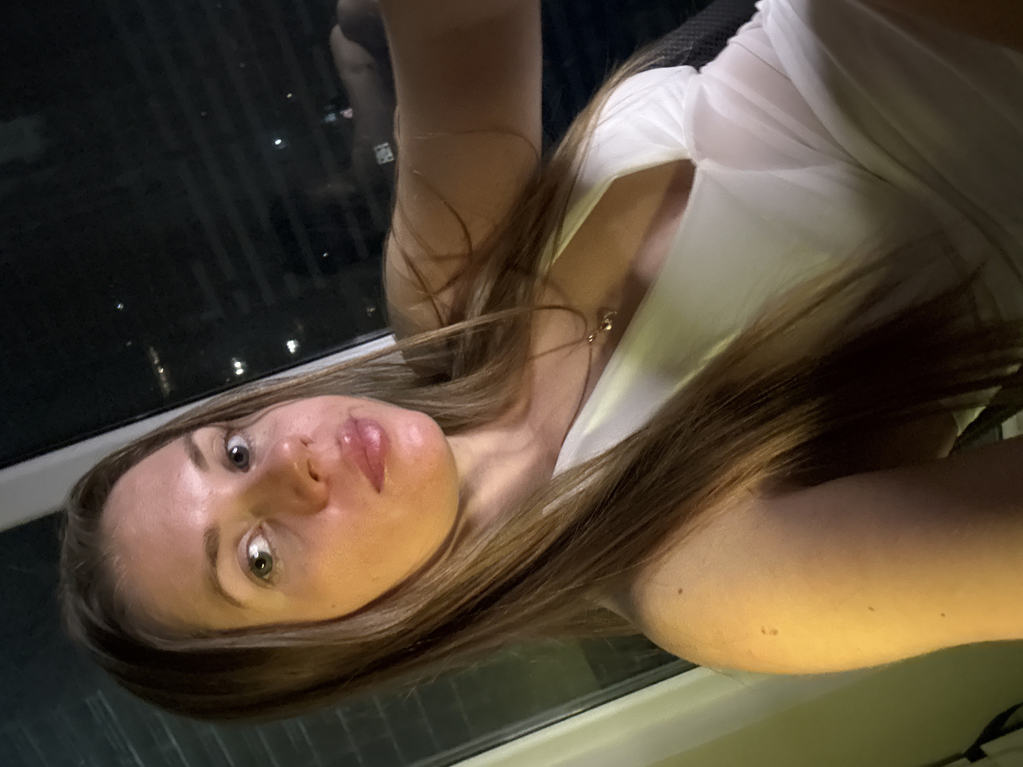 A young woman with long, light brown hair is depicted in a close-up, eye-level shot, appearing to be reclining indoors. Her face, framed by her hair, looks directly at the viewer with wide, light-colored eyes and full, glossy lips. She wears a white or cream-colored top featuring soft, gathered fabric around the neckline, and a delicate gold-toned necklace with a small pendant is visible on her chest.
The background is dark, indicating nighttime, with faint, scattered lights of an urban landscape visible through a window or glass partition. Reflections on the glass hint at the interior setting. A warm, yellowish light illuminates the woman's face and upper body from the right side, creating highlights and a soft glow. Her expression is calm and direct. The scene suggests a quiet moment in a high-rise building or apartment overlooking a city at night, specifically in Batumi, Georgia. No text is visible in the image.