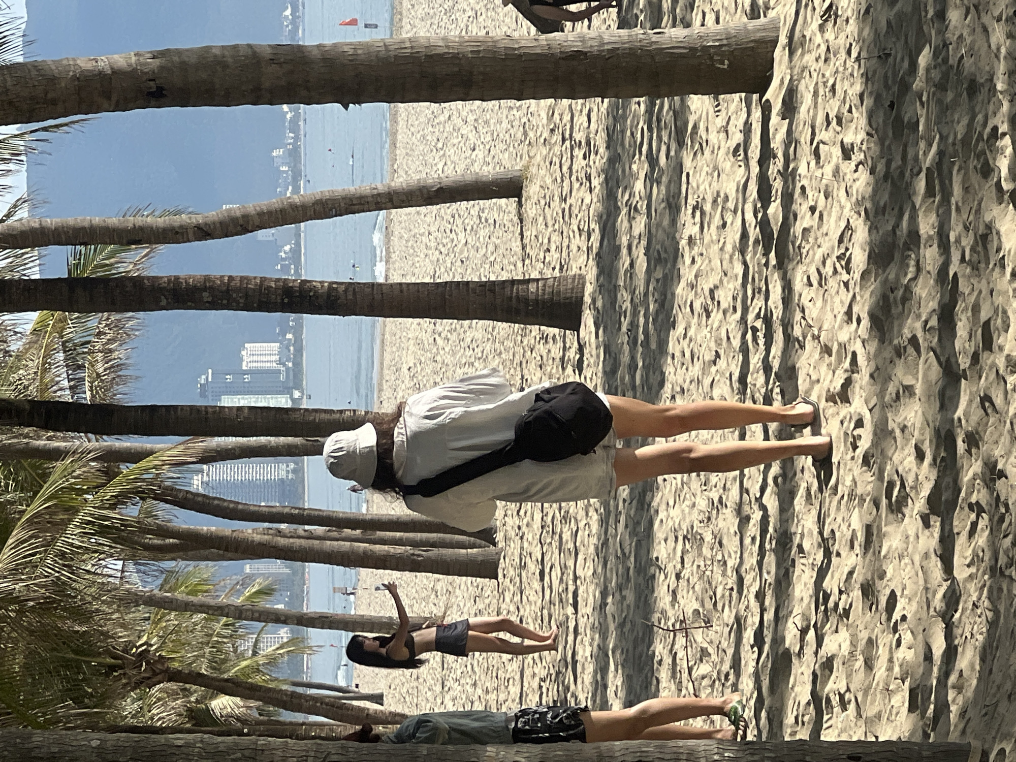 The image captures a scene on a sandy beach in Đà Nẵng, Vietnam, likely My Khe Beach, characterized by its numerous palm trees lining the shore and the clear blue ocean in the background. In the mid-ground, two women are visible. One, facing away from the camera, wears a white shirt, shorts, and a bucket hat, carrying a black bag across her chest. She appears to be taking a photo or video with her phone. To her left, a second woman, also facing away, is dressed in a bikini and a dark skirt, holding a smartphone and posing for a selfie. Further back, on the beach and in the ocean, several other individuals are present, enjoying the water and the sun. The city skyline, featuring modern high-rise buildings, is visible in the distance beyond the ocean, indicating an urban coastal setting. The foreground is dominated by the textured sand and the shadows cast by the palm trees.
