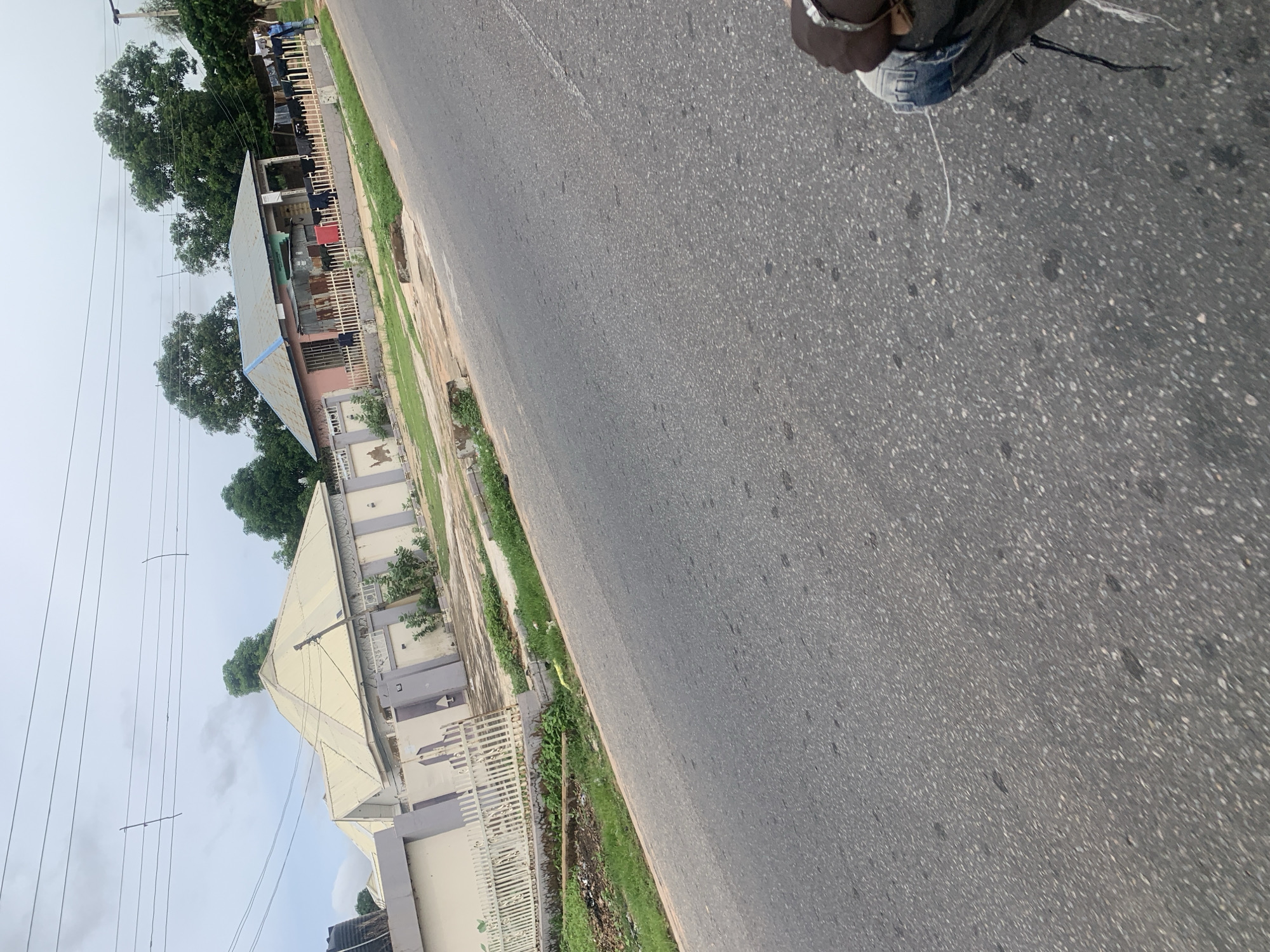 The image depicts a street scene in Jalingo, Nigeria. On the left side of the asphalt road, a pale yellow building with grey accents and a white fence is visible. Beyond this structure, another building with a pitched roof and a light-colored facade can be seen. To the right of the road, a pink-colored building with a corrugated metal roof and a fence in front is present. Laundry is hung on a clothesline in front of this building, and a person in a blue shirt stands near it. Tall green trees form a backdrop to the buildings. The road surface is asphalt with some visible debris. In the foreground, a person's hand wearing a bracelet and a portion of their leg in ripped jeans are visible, suggesting the photo was taken from a moving vehicle. The sky is overcast with a few wispy clouds.