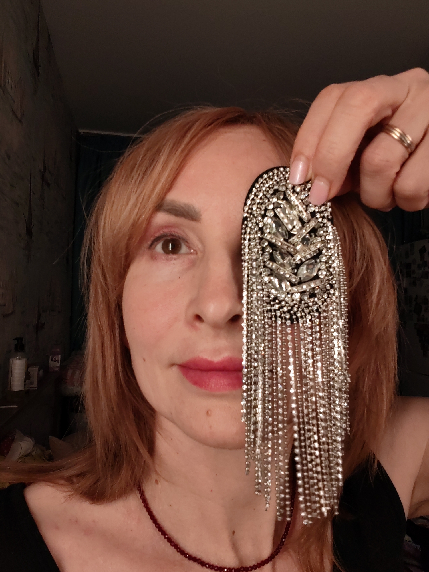 A woman with light reddish-brown hair and a neutral expression is captured in a close-up, chest-up shot, looking directly at the camera. She holds a large, ornate, black decorative piece adorned with sparkling silver rhinestones and long, dangling chains of smaller rhinestones, obscuring her right eye. Her visible left eye is brown, and she wears pink lipstick.
She is dressed in a dark top, with a red beaded necklace visible around her neck. On her right hand, which holds the accessory, a simple gold band is seen on her ring finger. The background on the left features a dark, textured wall, possibly patterned wallpaper, beneath which are a pump-dispenser bottle and other indistinct objects on a surface. The remainder of the background is dark and out of focus, indicating an indoor setting, likely a home. The lighting suggests an interior with artificial light, possibly evening. No discernible text is visible. The image conveys a playful or fashion-oriented pose, showcasing the elaborate accessory.