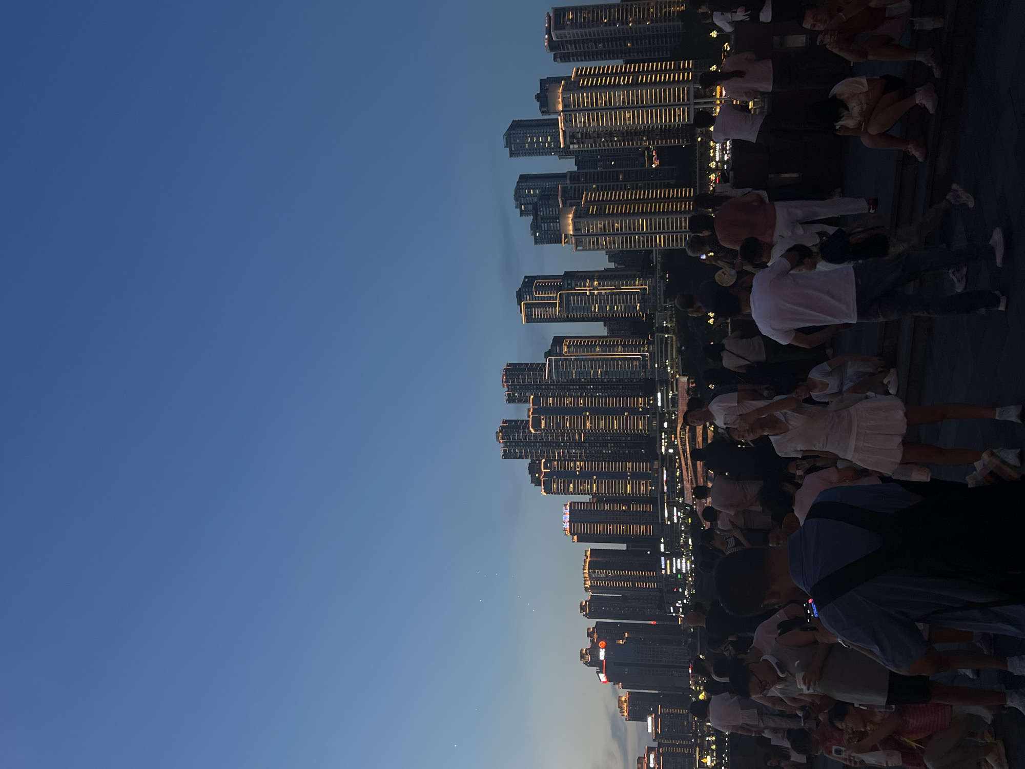 The image depicts a nighttime cityscape with numerous illuminated skyscrapers in the background, suggesting an urban environment. In the foreground and midground, a large crowd of people are gathered. The individuals are diverse in age and are engaged in various activities, including standing, sitting, and holding mobile phones, indicating they are likely observing something or socializing. The sky is a deep blue, transitioning to a lighter hue near the horizon, typical of dusk or early night. The architecture of the buildings features a modern, vertical design with many windows, some of which are lit, adding to the city's glow. The overall scene suggests a public gathering or event taking place in a well-developed city, possibly a waterfront area given the unobstructed view of the skyline. The location is identified as Yuzhong District, China.
