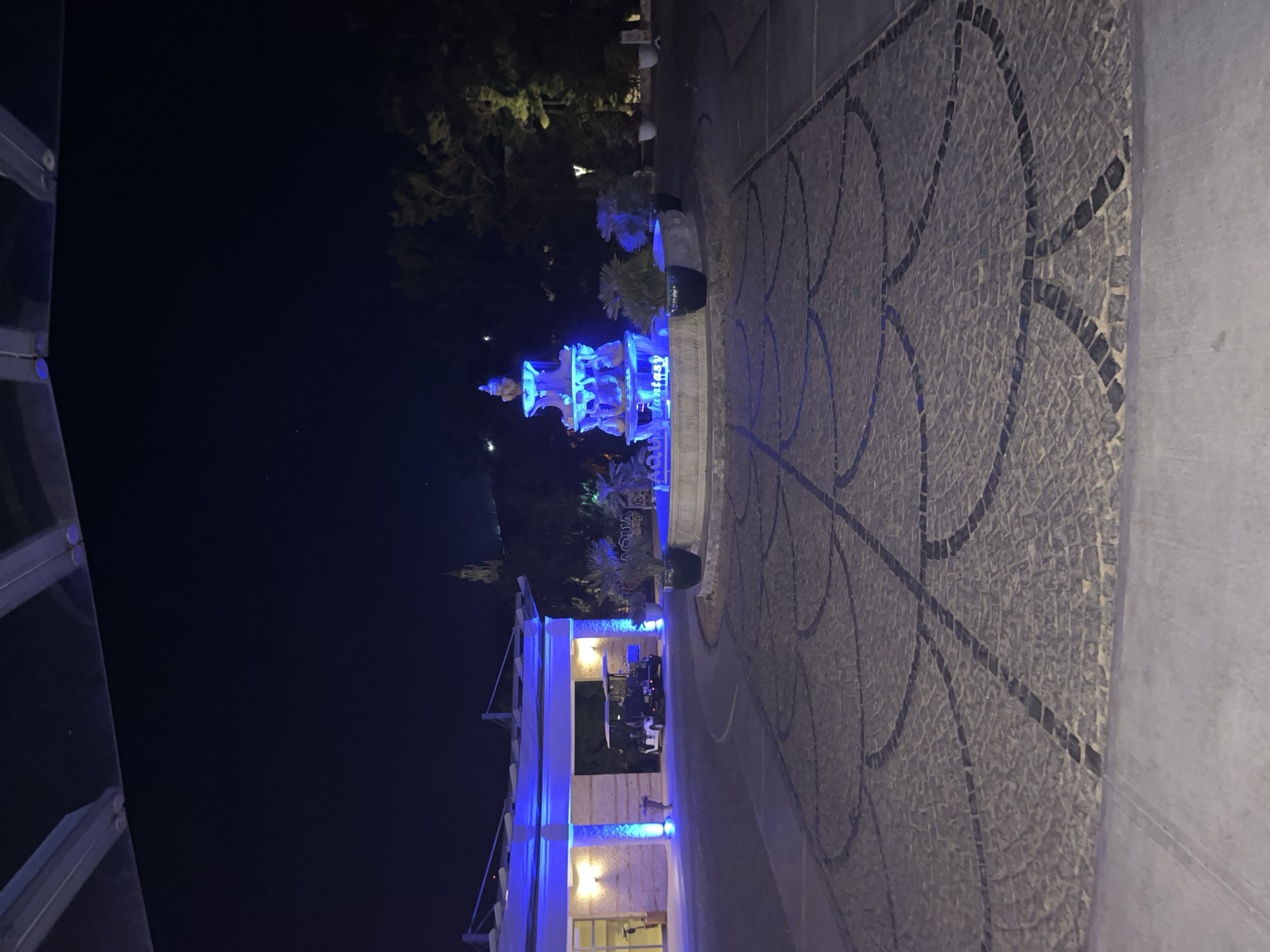 This media file captures an outdoor nighttime scene, likely within a resort or public leisure area in Selçuk, Turkey. The time of day is clearly night, evidenced by the dark sky and the reliance on artificial illumination.
The central subject is a multi-tiered fountain, prominently lit with a striking blue light, making it the focal point of the image. The words "Fantasy" and "Aqua" are visible on the fountain's structure. To the right of the fountain, a decorative pathway or plaza is visible, paved with small stones forming an intricate, swirling mosaic pattern.
On the left side of the frame, a building with architectural elements like columns and arches is partially visible, also illuminated with blue light. Inside or under a covered section of this building, several indistinct figures can be seen seated at tables, suggesting an active dining or social area. A golf cart is parked nearby, indicating a larger complex where such transport might be used.
In the background, dark foliage from trees can be discerned. Further in the distance, on a hilltop, a faint, illuminated structure, possibly a historical building or a castle, adds a geographical context to the scene. There are no discernible weather conditions other than darkness. The overall mood conveyed by the blue lighting is calm and atmospheric.